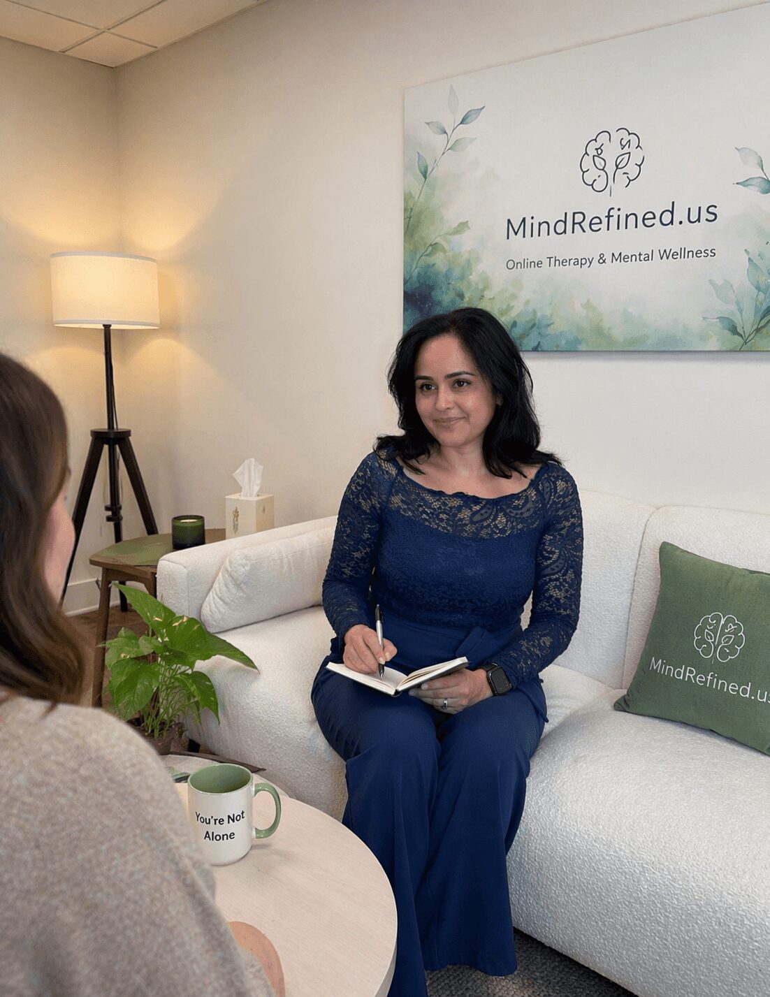 Woman sitting on a white sofa, holding a notebook, in a therapy office with a sign on the wall.