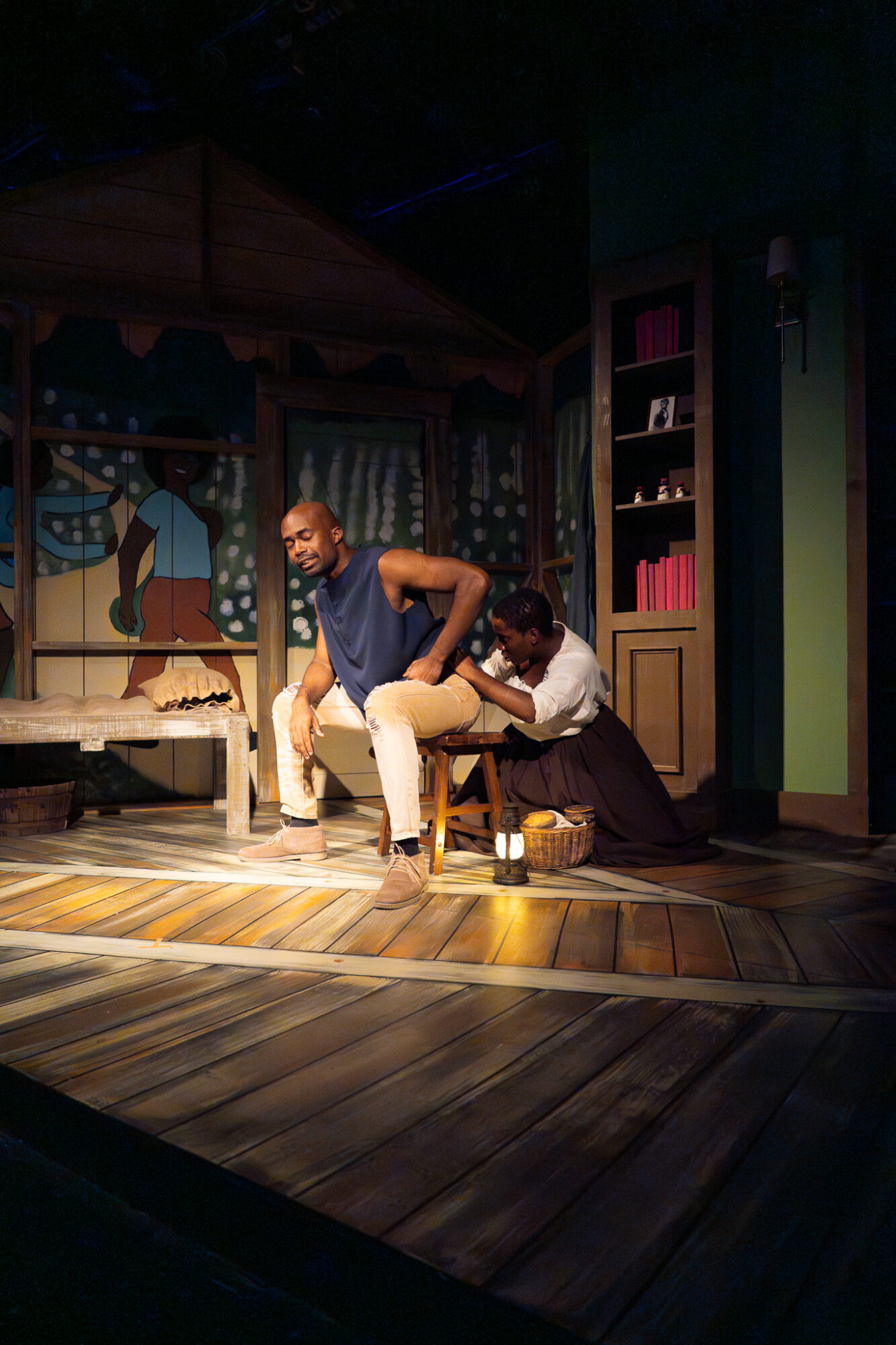 Two people on stage, one seated and one kneeling, in a colorful room with shelves and a painted backdrop.
