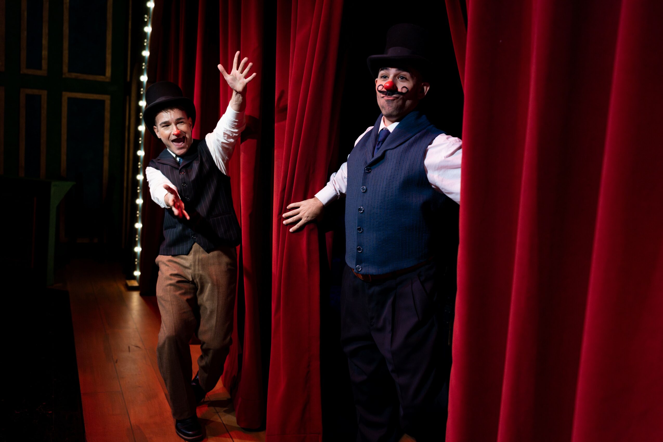 Two performers on stage with red curtains, one waving and the other smiling, both in costumes and makeup.