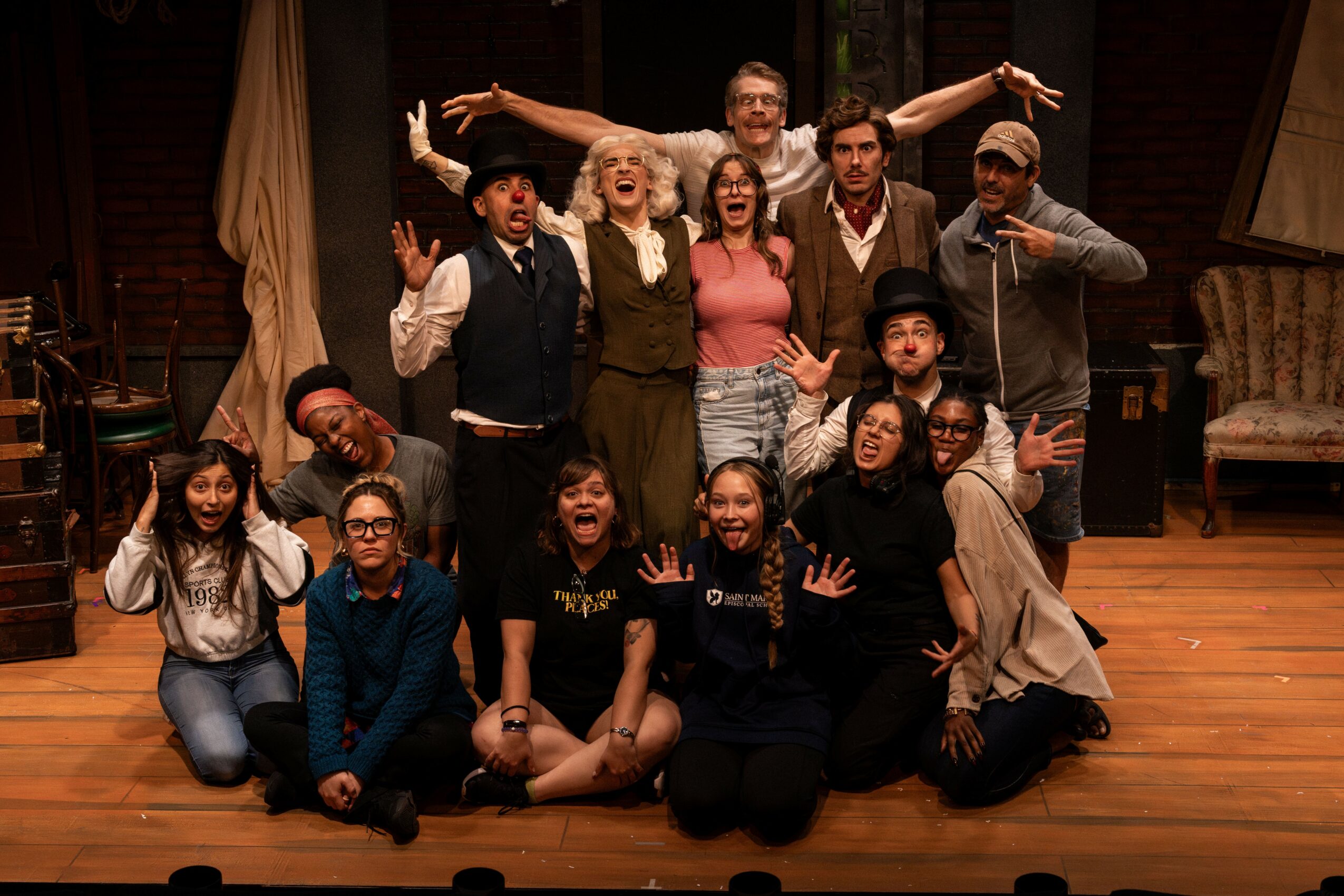 Group of people on stage, smiling and posing with arms raised, in a theatrical setting with wooden floor and furniture.
