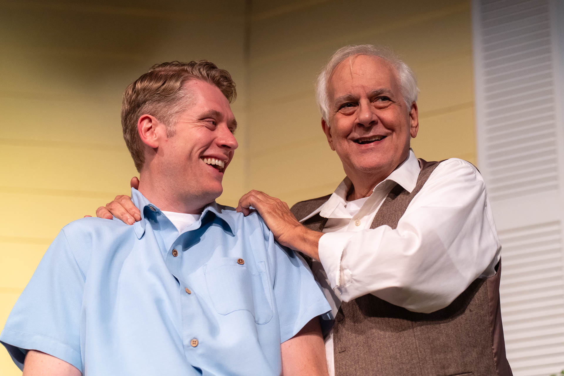 Two men smiling and talking, one with hand on the other's shoulder, in a room with yellow and white walls.