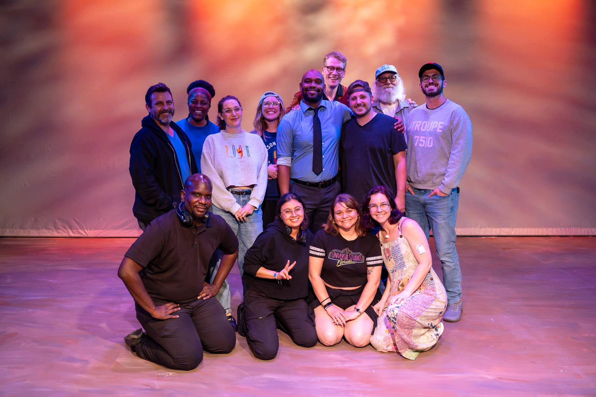 Group of twelve people posing on stage with colorful background, some standing and some kneeling, smiling at camera.