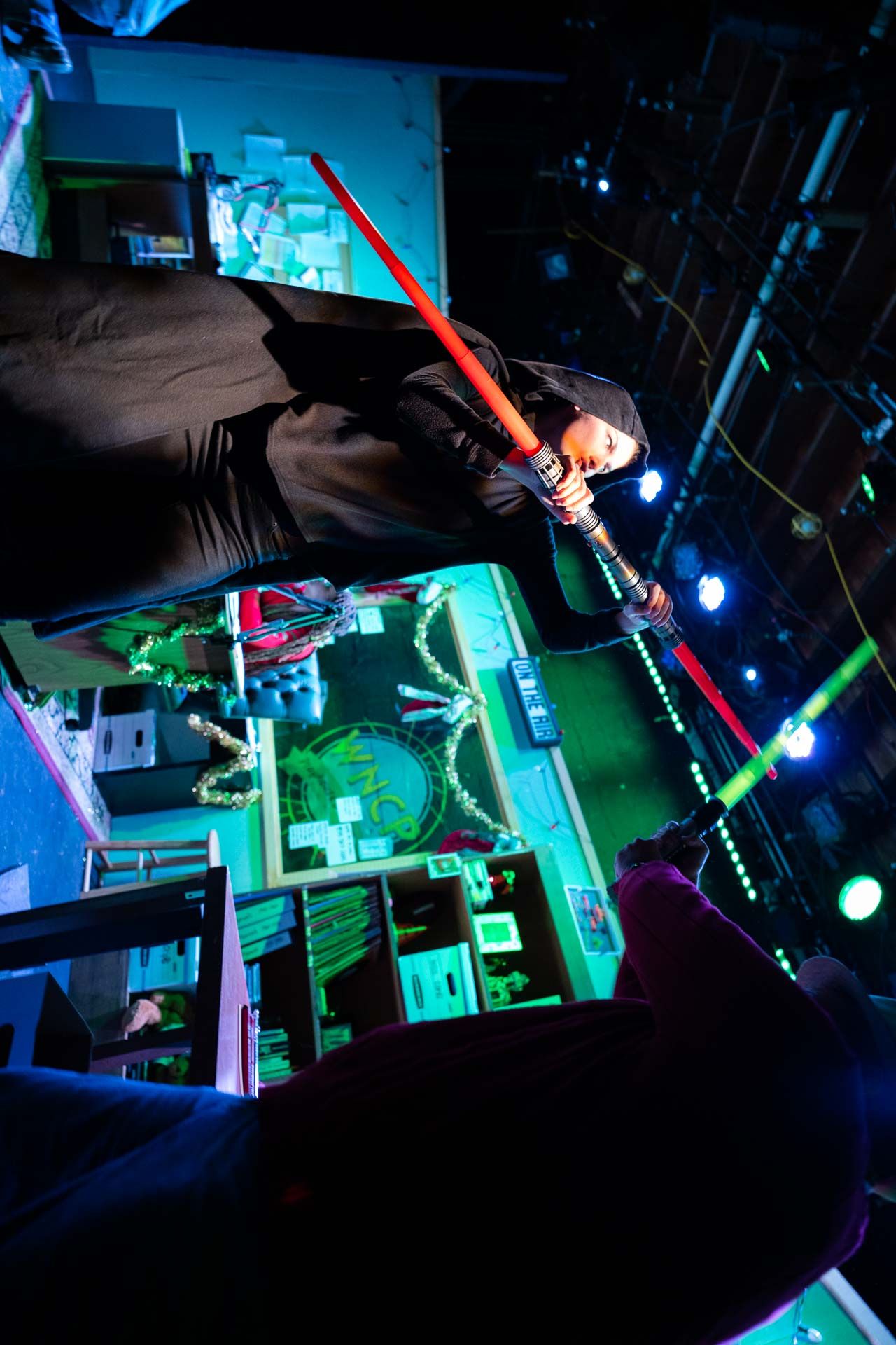 Person holding a red stick with a glowing green tip, standing in a room with colorful lights and shelves.