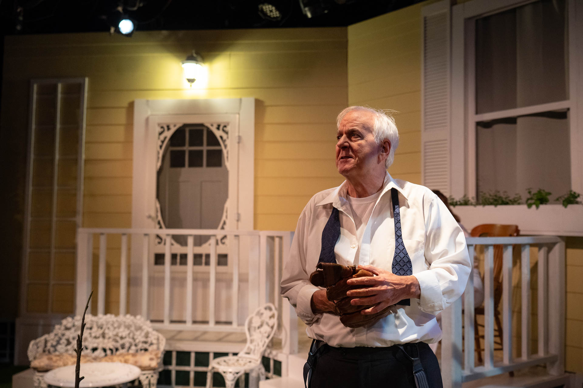 Older man with white hair holding a book, standing on a porch with yellow house background, white railing, and window.