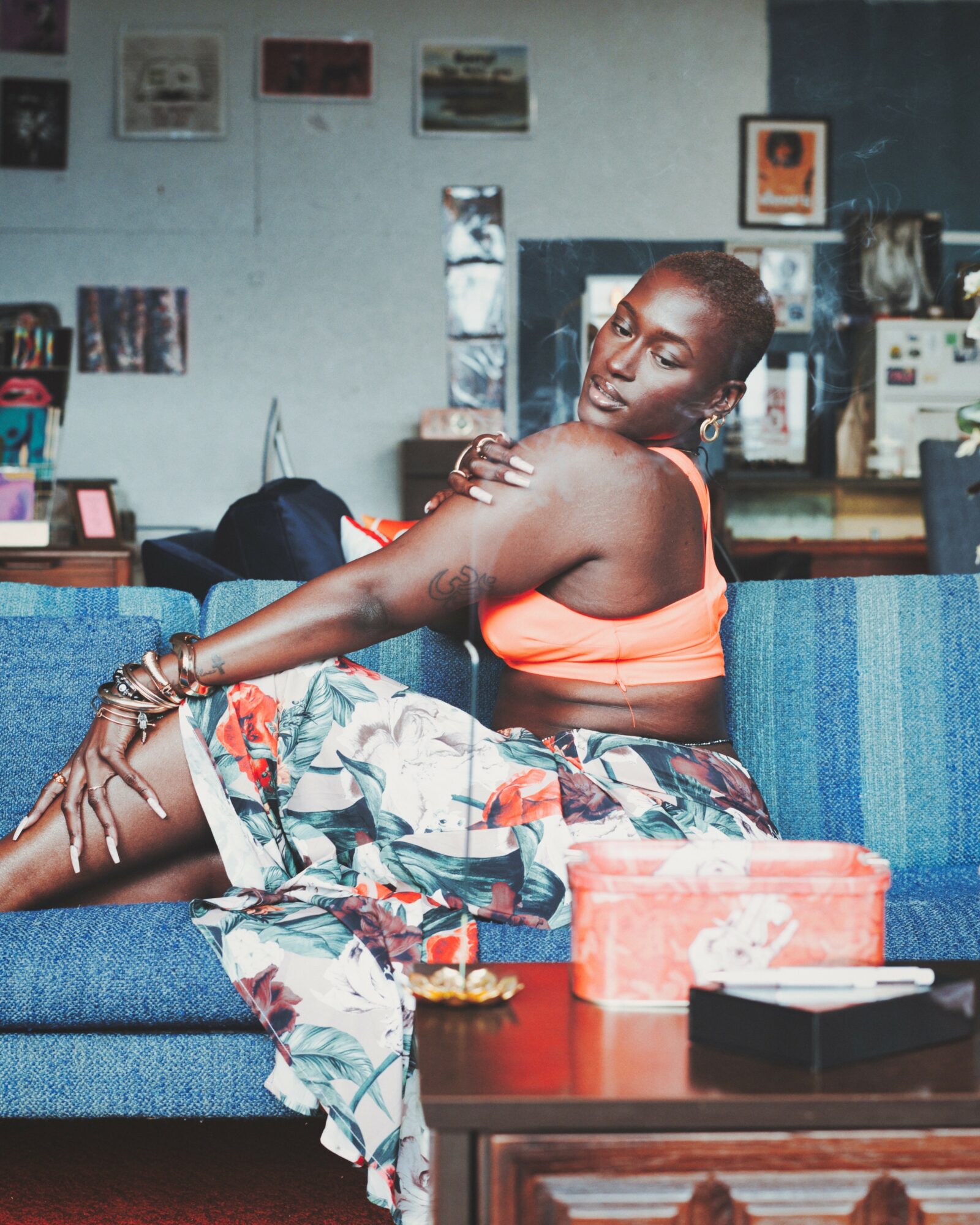 Woman sitting on a blue couch, wearing a coral top and floral skirt, in a room with pictures and objects on the wall.