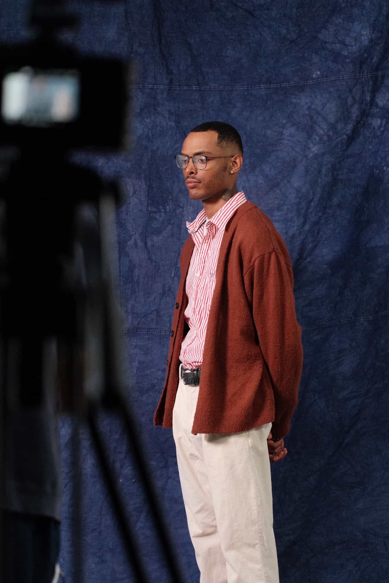 Young man with glasses wearing a pink striped shirt, brown cardigan, and beige pants, standing against a blue backdrop.