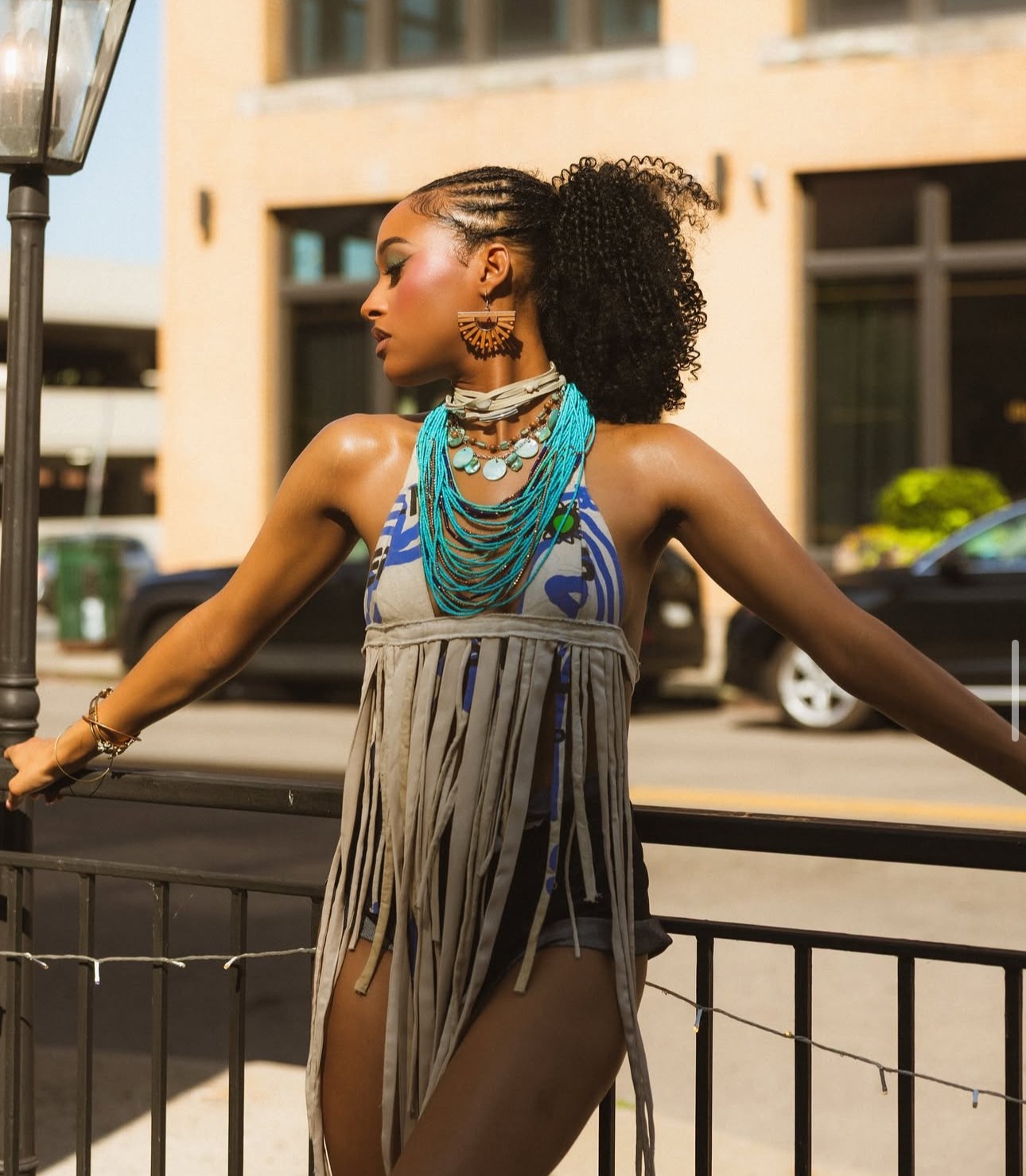 Woman with curly hair wearing layered necklaces and a fringed dress, standing outdoors near a railing.