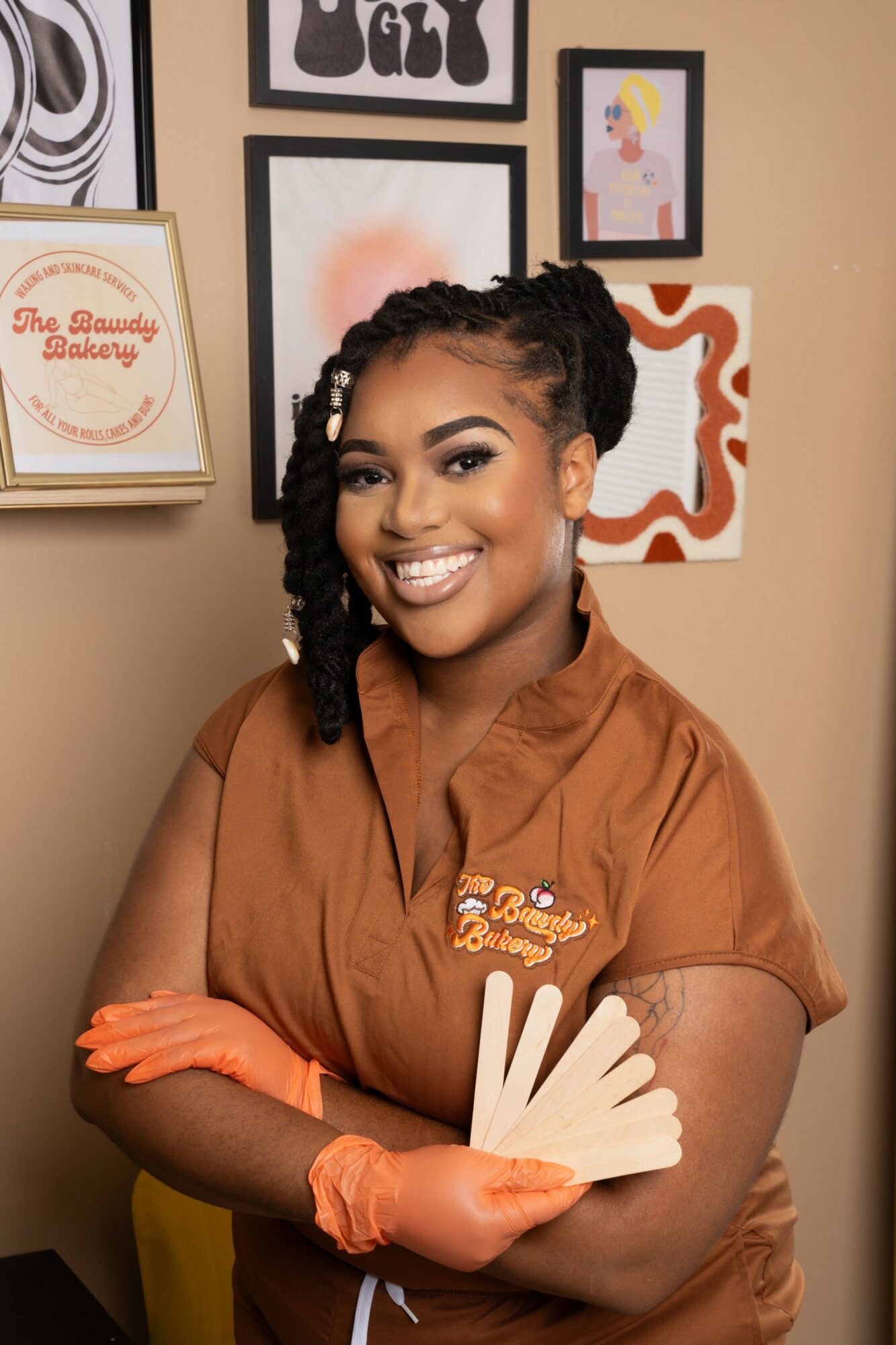 Smiling woman with braided hair wearing a brown uniform and orange gloves, holding color swatches in front of a wall with framed art.
