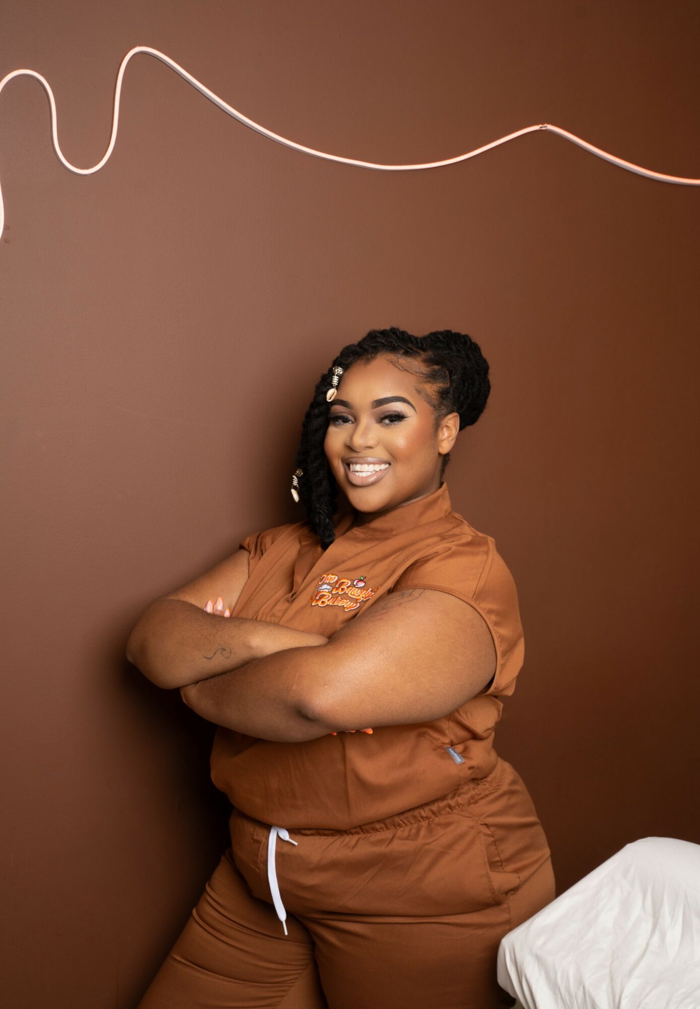 Smiling woman with arms crossed, wearing a brown outfit, standing against a brown wall with a white line decoration.
