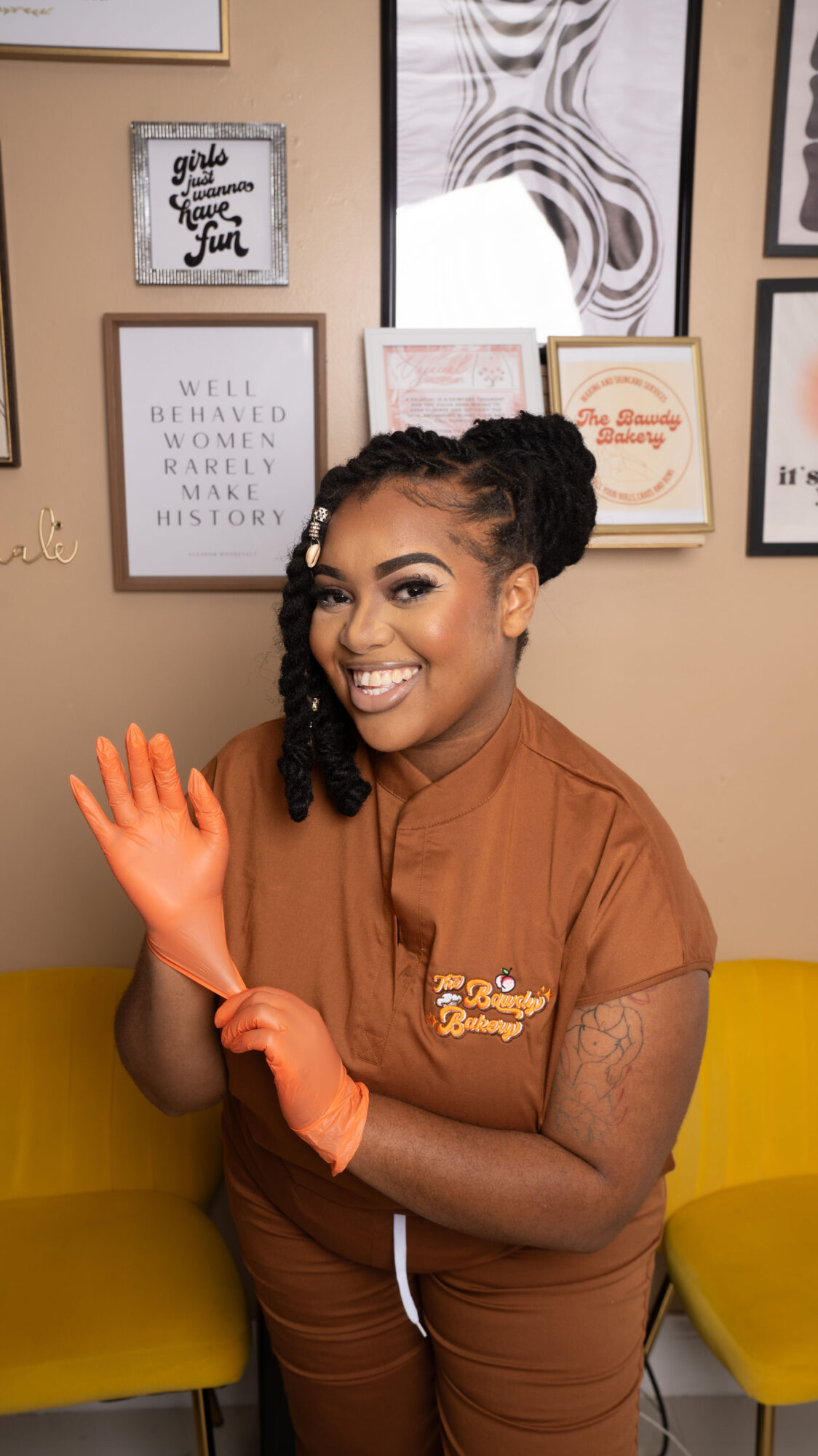 Smiling woman in brown uniform with orange gloves, standing indoors with framed pictures on wall behind her.