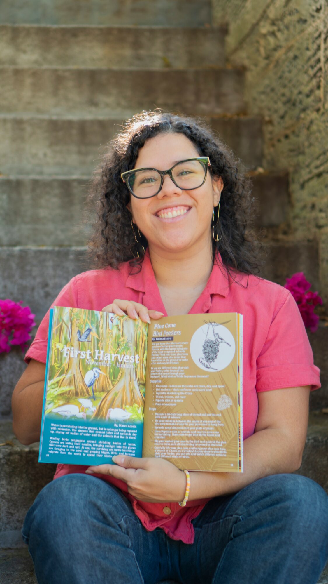 Woman with glasses and curly hair smiling, holding a colorful book outdoors with stone stairs behind her.