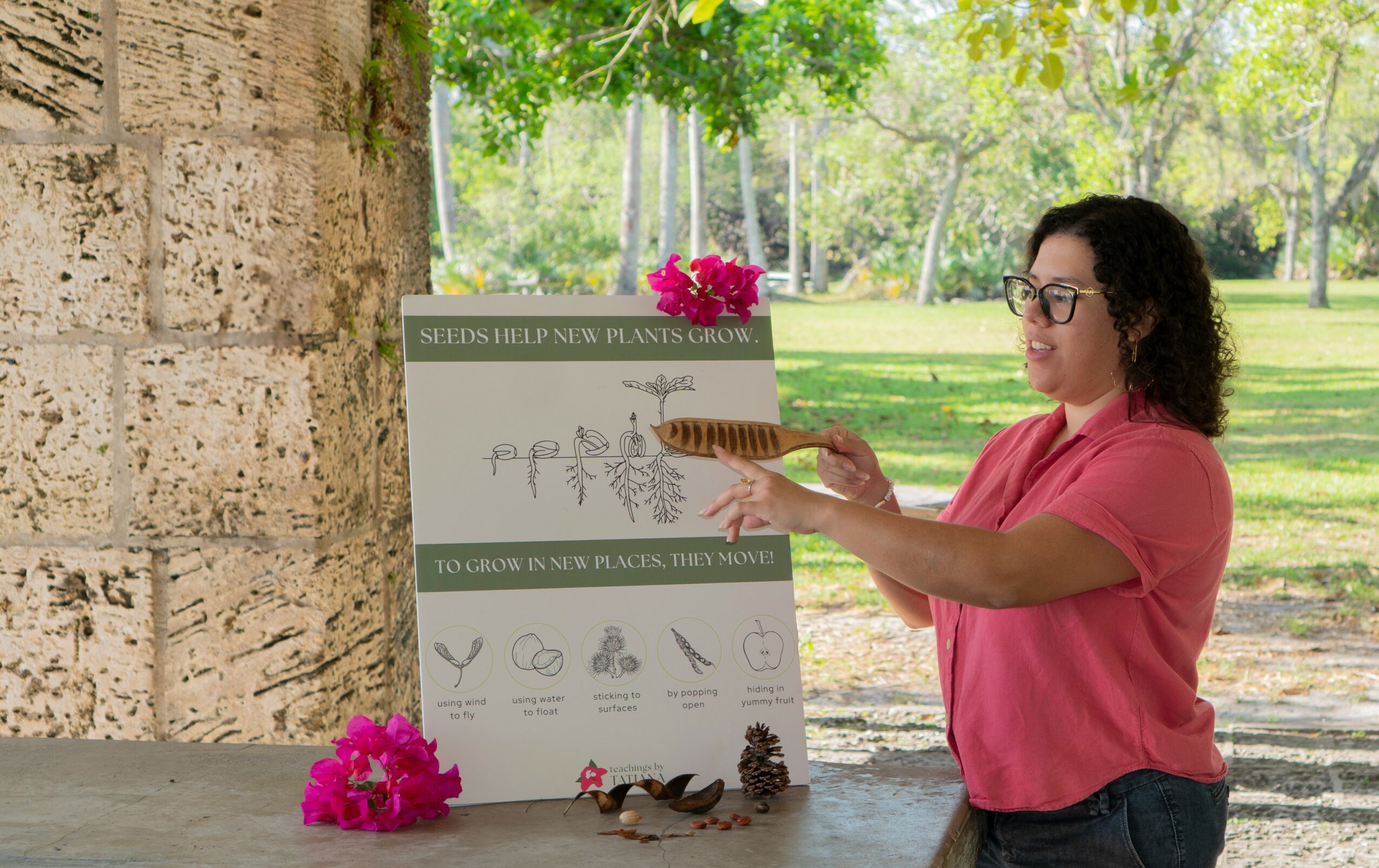 Woman with glasses in pink shirt pointing at educational poster outdoors with trees and grass.