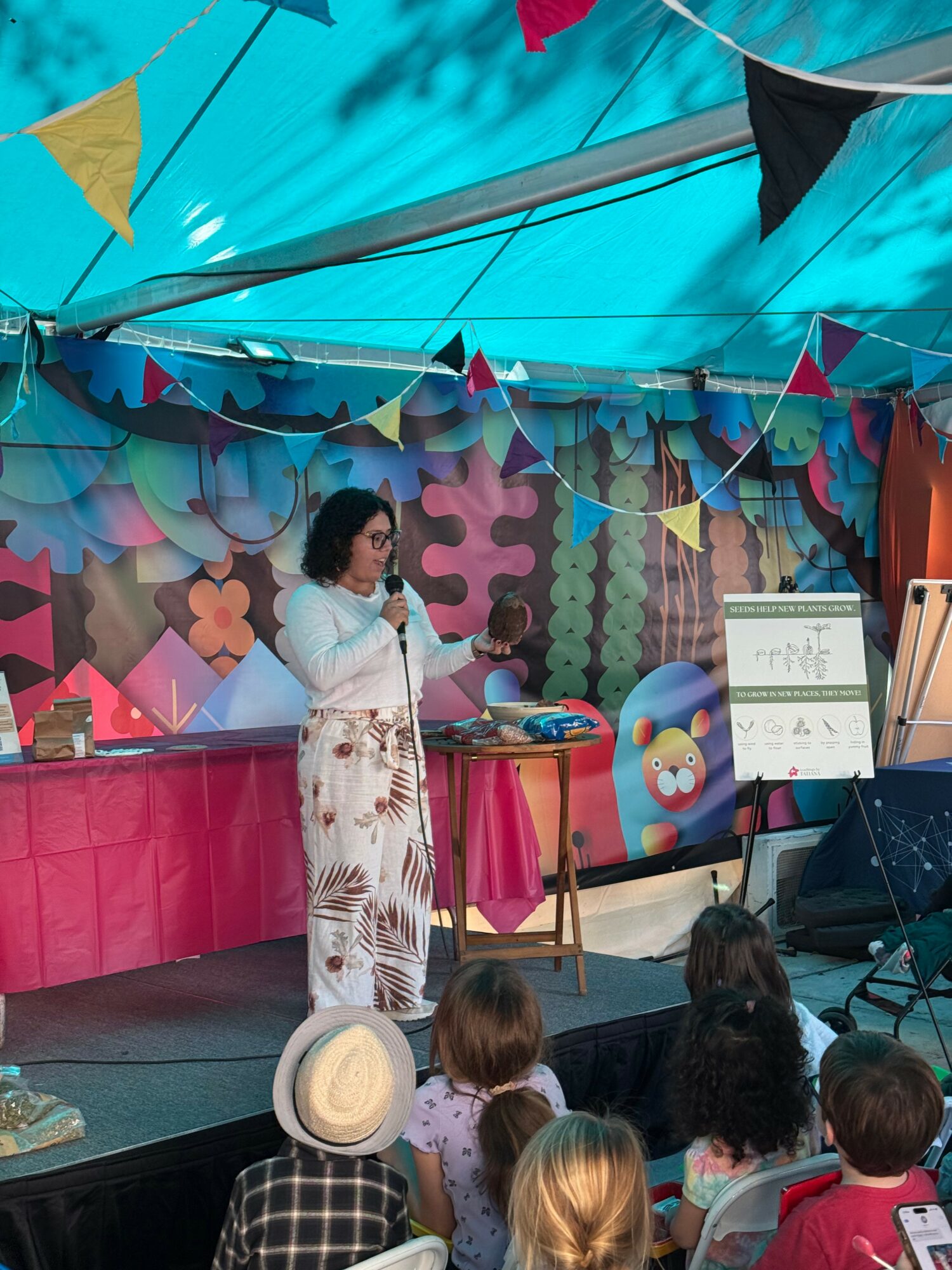 Woman speaking on stage with children seated in front, colorful decorations and a whiteboard behind her.