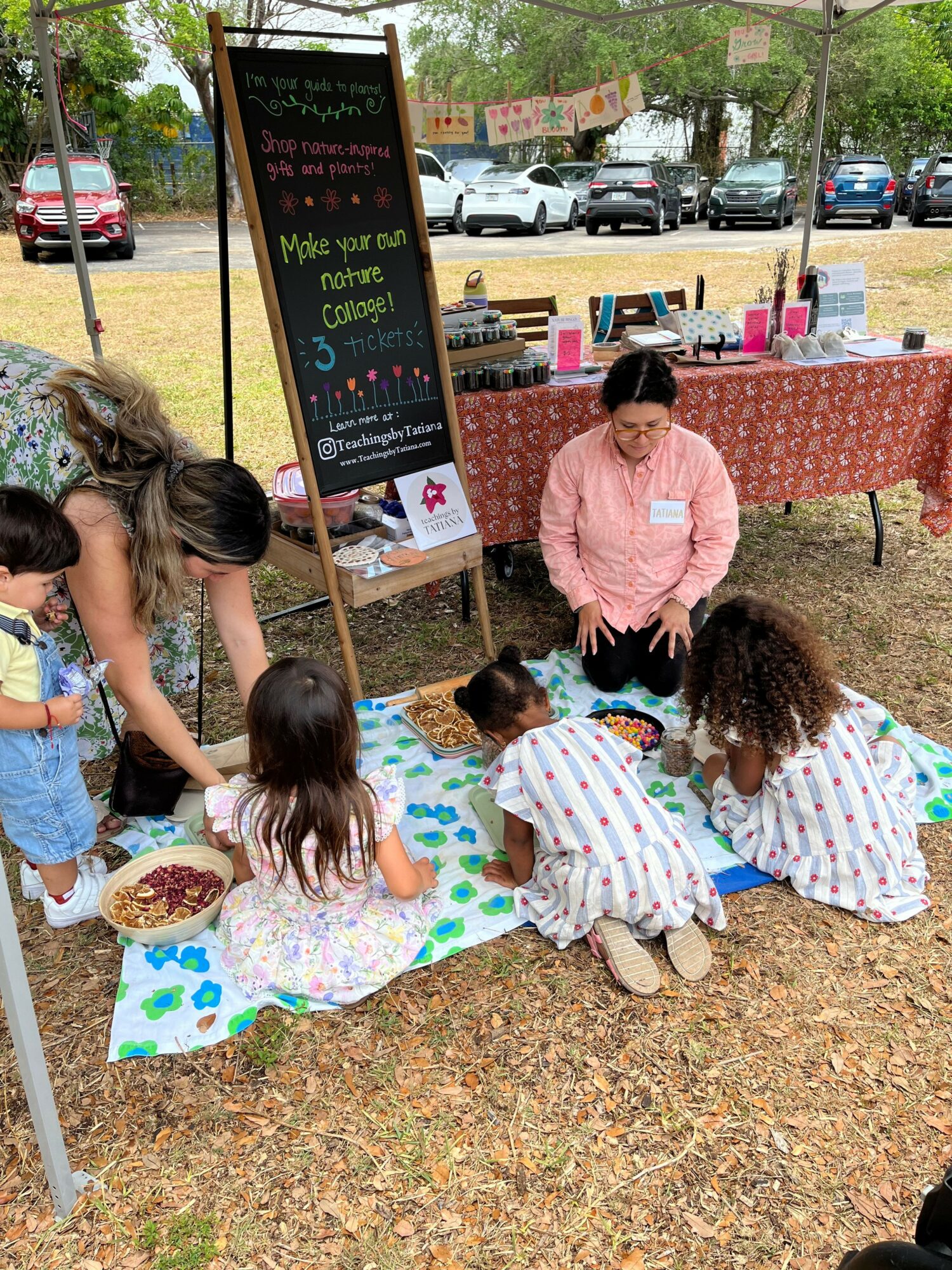 Children sitting on a colorful mat with an adult woman at an outdoor event, with tables and a chalkboard sign nearby.