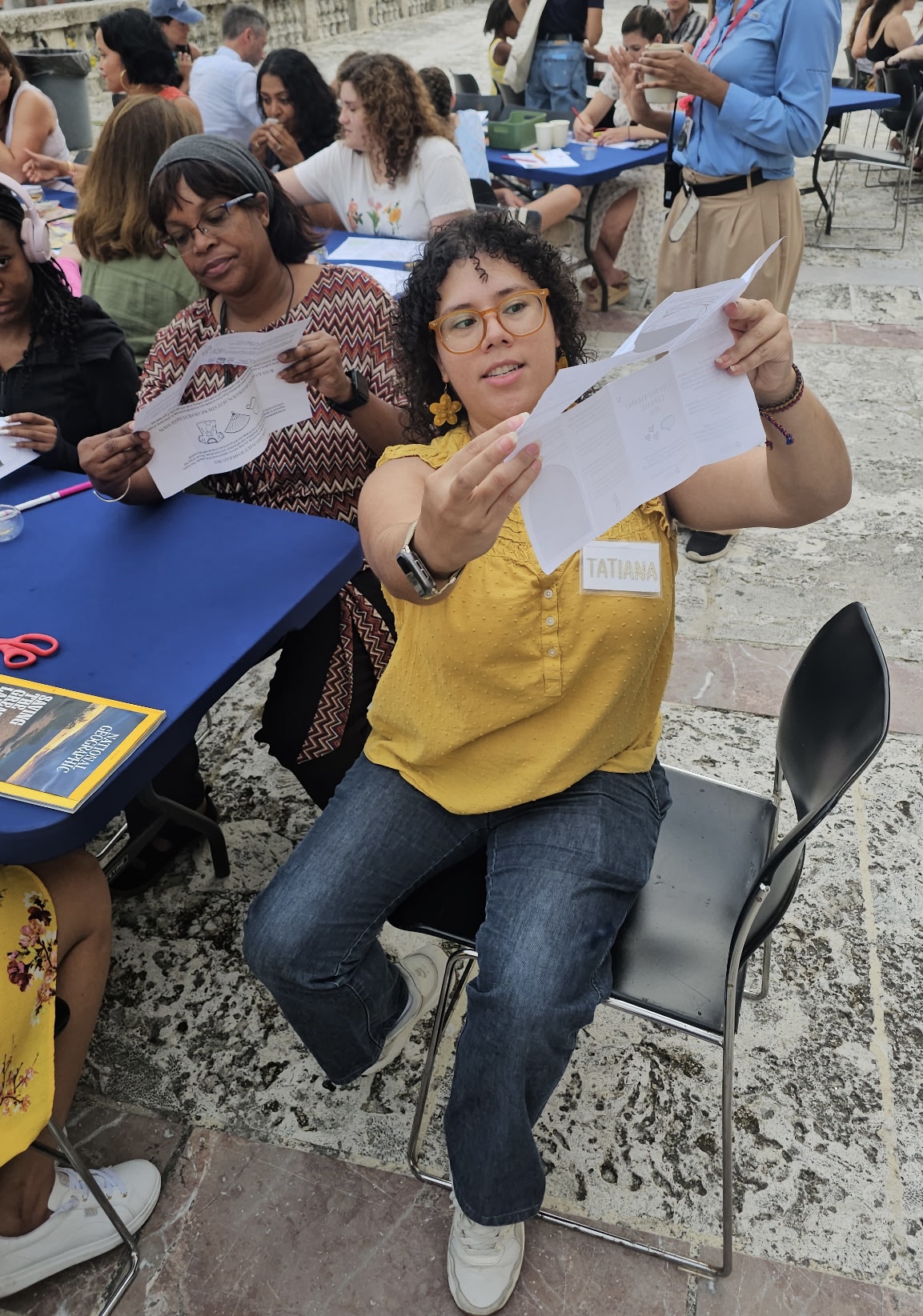 Woman in yellow shirt sitting on a chair, holding and looking at a paper, surrounded by people at an outdoor event.