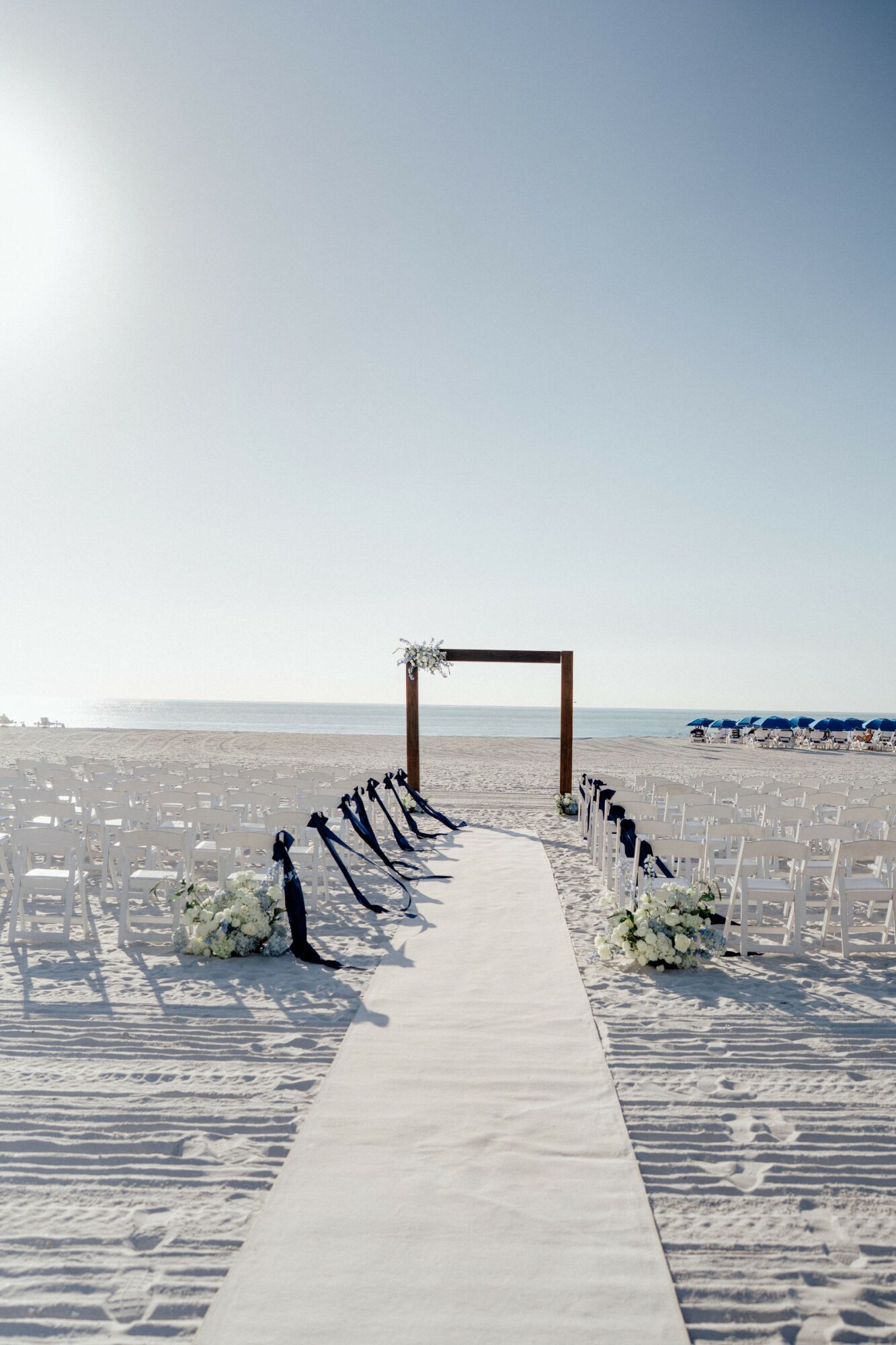 Beach scene with a white aisle leading to a wooden arch, decorated with flowers, under a clear sky.