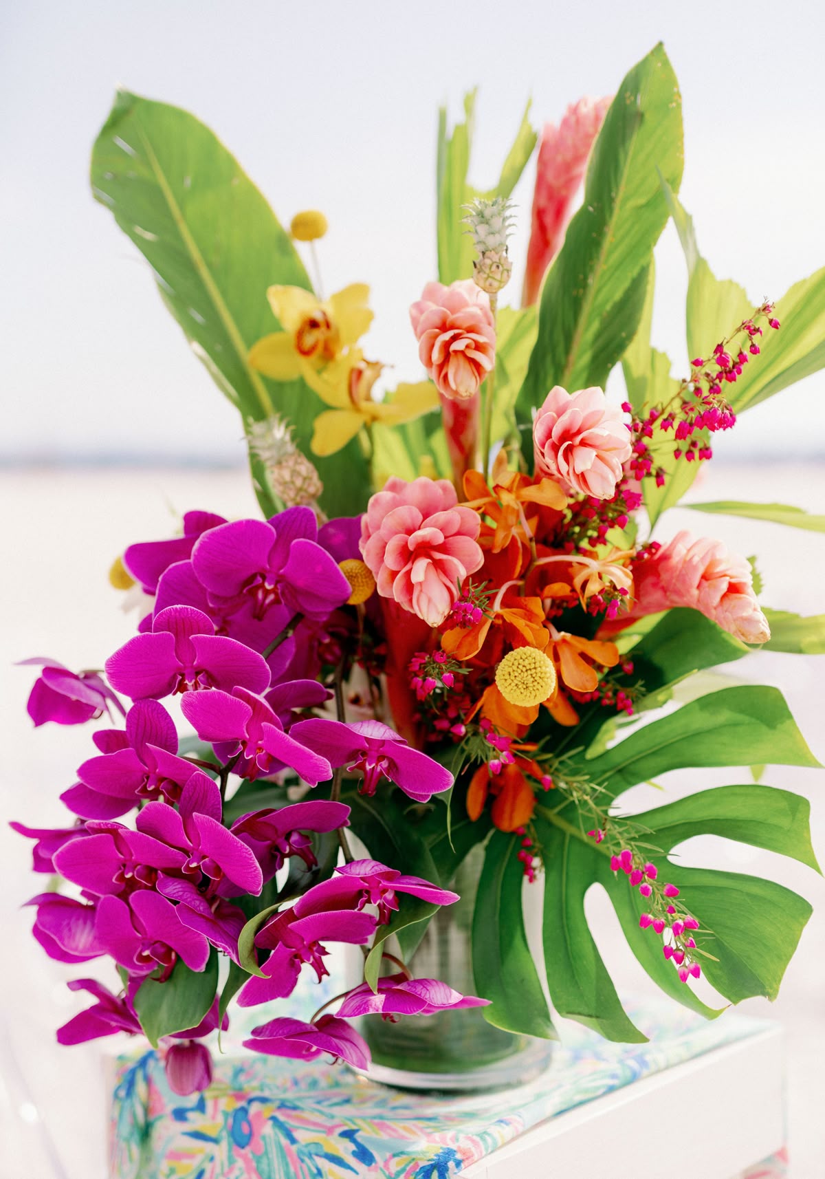 Colorful flower arrangement with pink, yellow, and orange flowers and large green leaves in a glass vase.