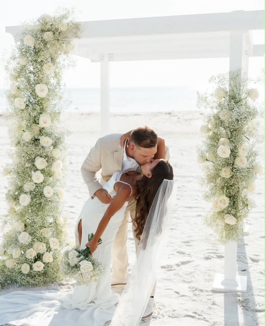 Couple kissing under decorated arch on beach, with ocean in background, floral arrangements on sides.
