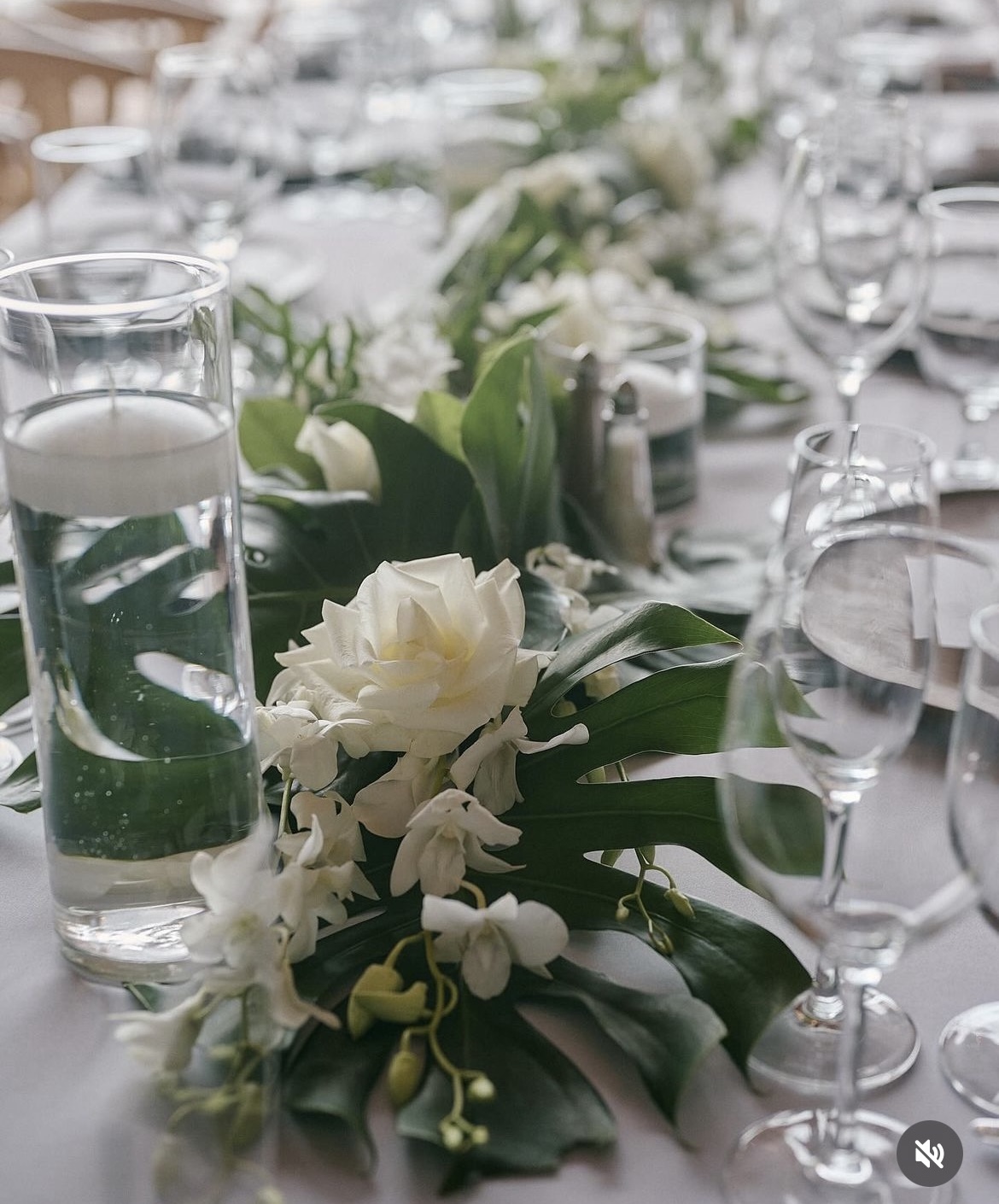 Elegant table setting with a floral centerpiece of white flowers and green leaves, surrounded by glassware and tableware.