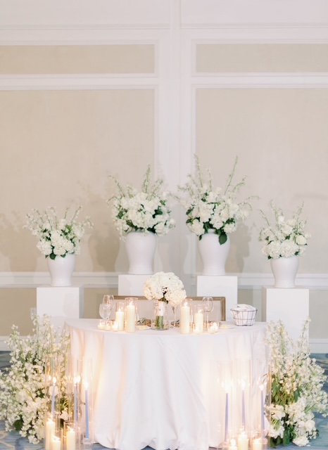 Table with white flowers and candles, three large flower arrangements in the background, in a bright room.