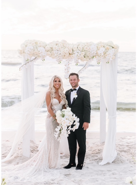 Bride and groom stand under floral arch on beach, holding hands, smiling, ocean in background.