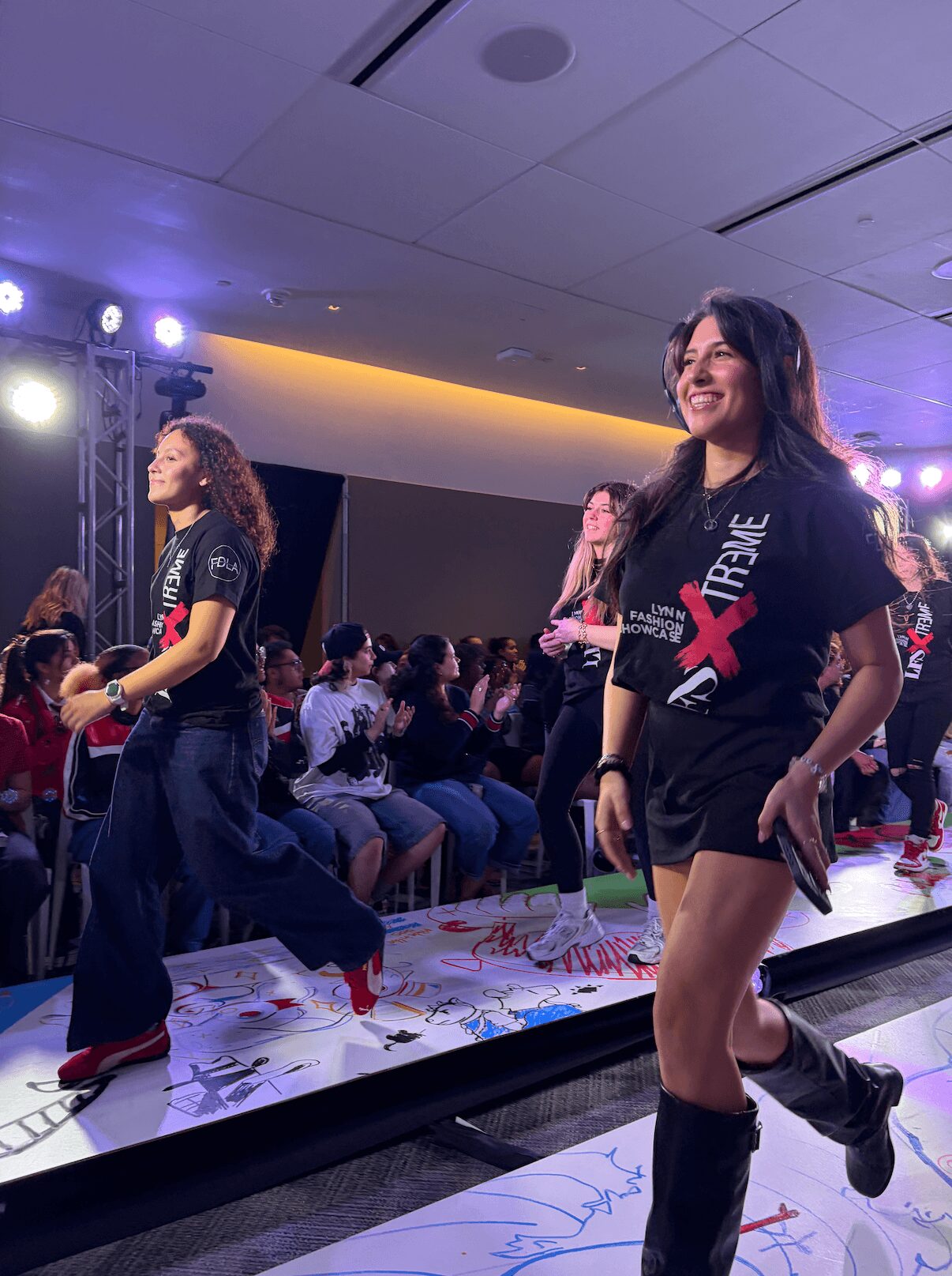 Two women walking on a runway during an event with an audience in the background.