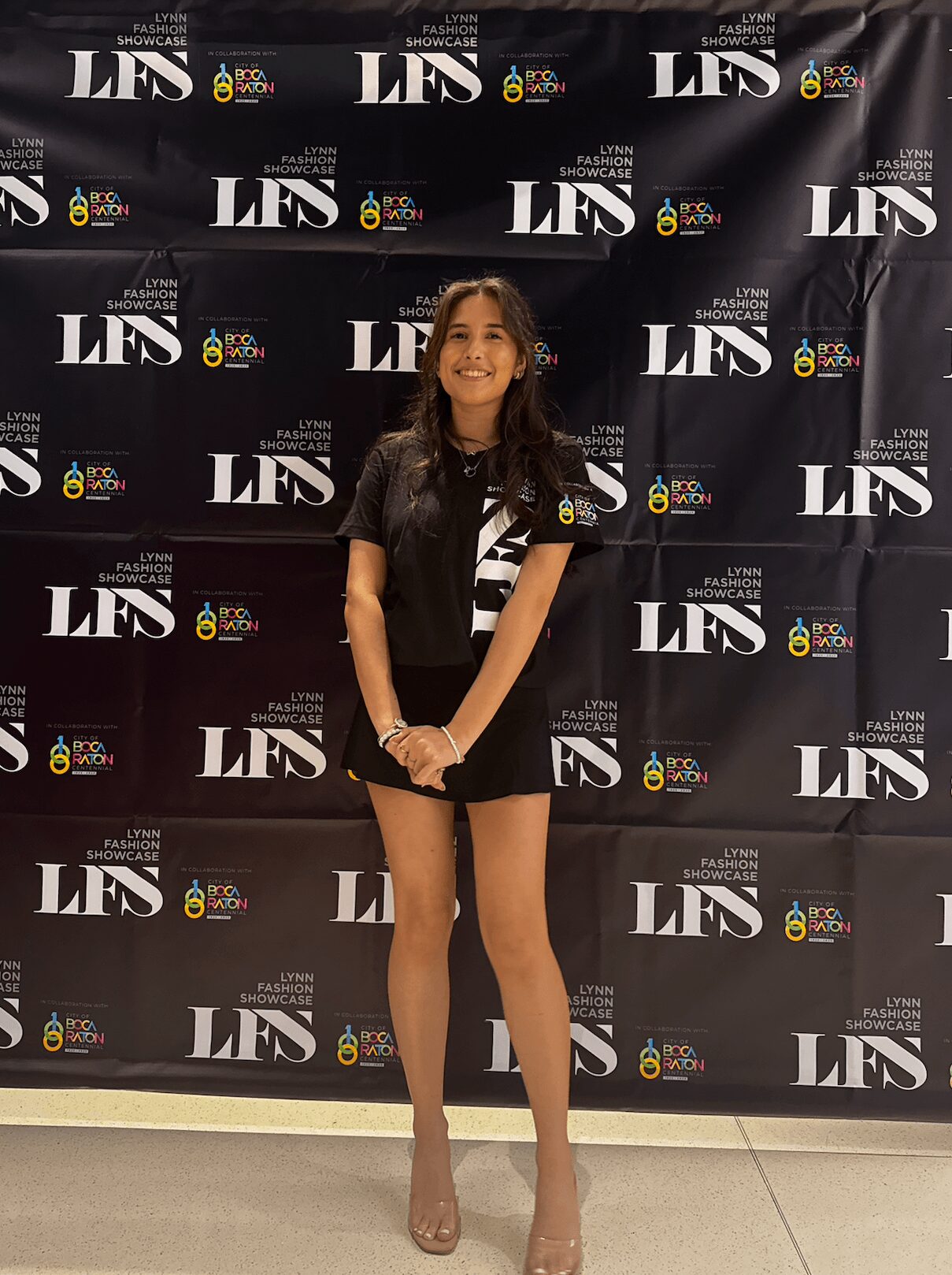Young woman standing in front of a black backdrop with logos, smiling, wearing a black blazer, striped shirt, and beige heels.