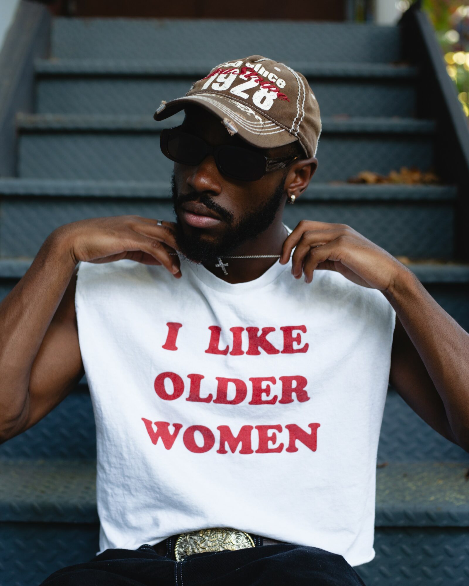 Person wearing a cap and sunglasses, holding a shirt with text, sitting on stairs outdoors.