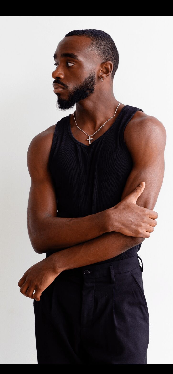 Young man with a beard and short hair, wearing a black sleeveless shirt and necklace, standing sideways with arms crossed.