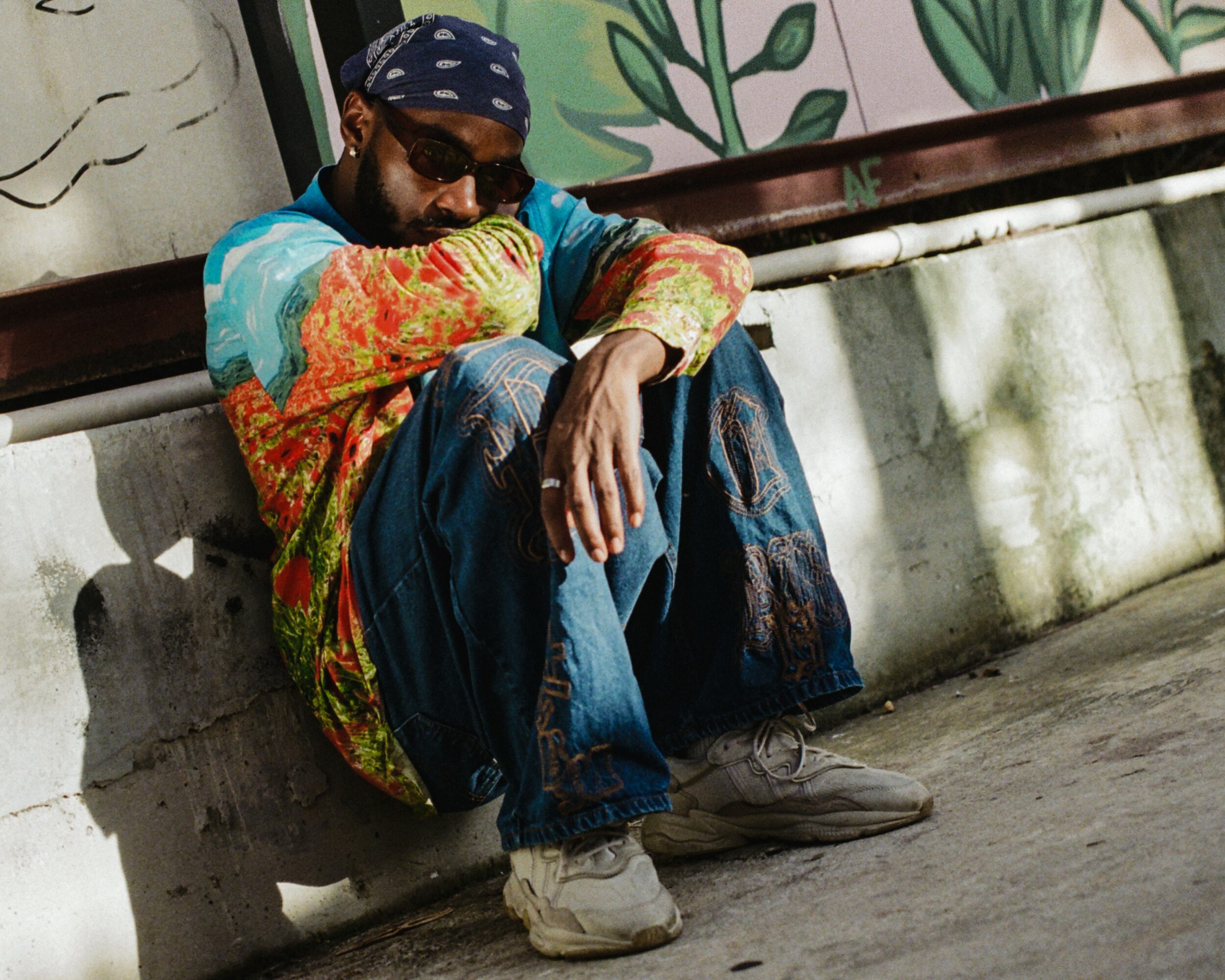 Person sitting on ground, wearing sunglasses, colorful jacket, jeans, and a bandana, leaning against a wall.