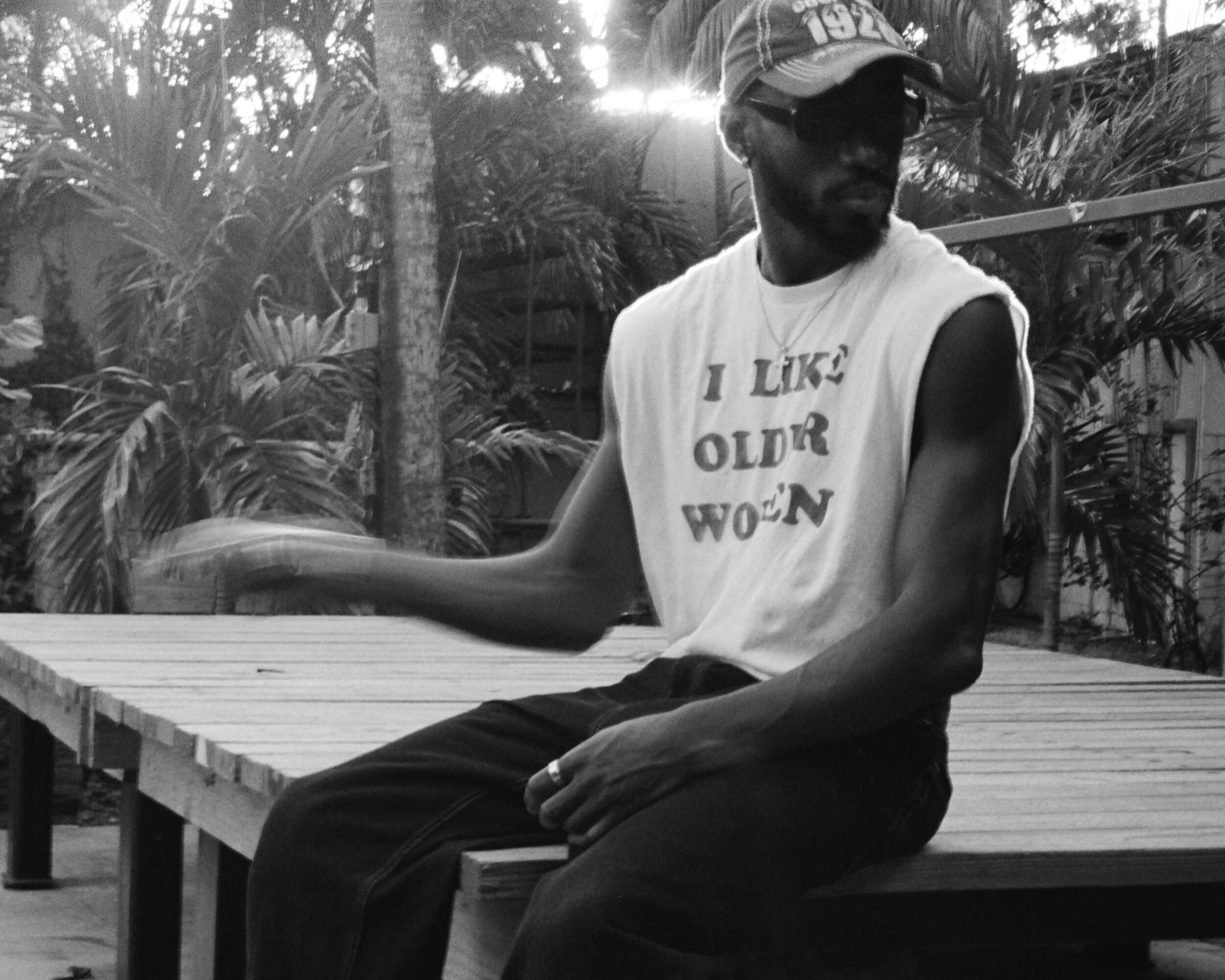 Person sitting on a wooden platform outdoors, wearing a cap and sleeveless shirt, with trees and plants in background.