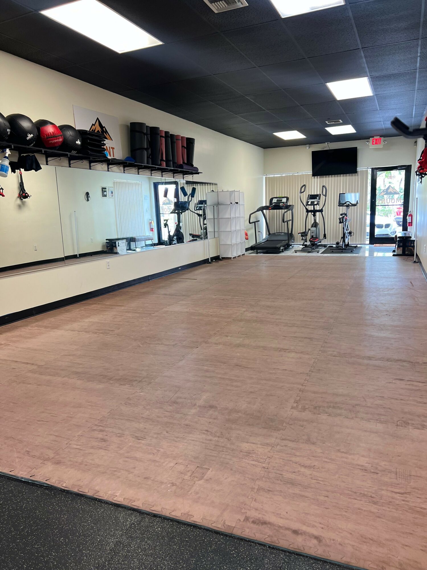 Empty gym with exercise equipment, mirrors, and a large open floor space, with a door and window in the background.