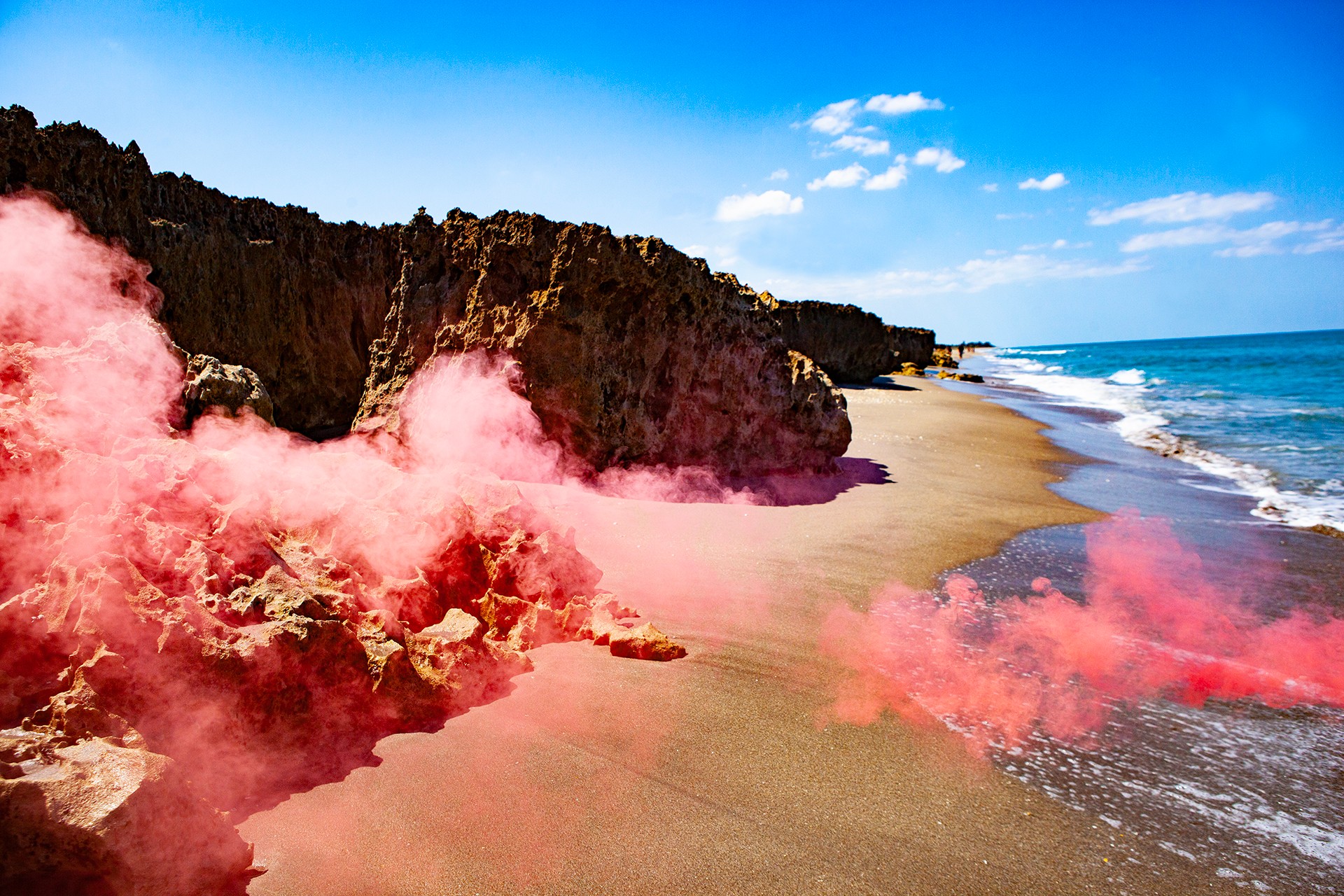 Beach with cliffs, ocean waves, and pink smoke or fog on sand, under a blue sky with clouds.