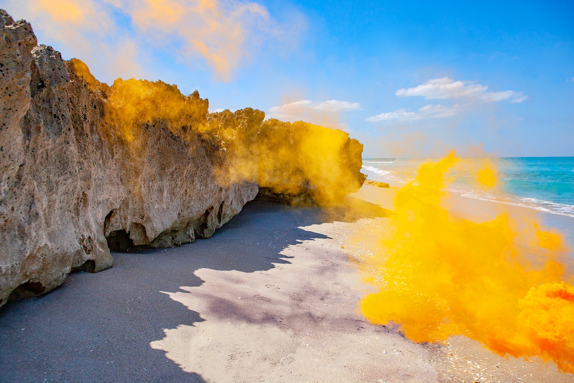 Cliffside beach with yellow smoke or fog near rocks, sandy shore, blue sky, and ocean in the background.