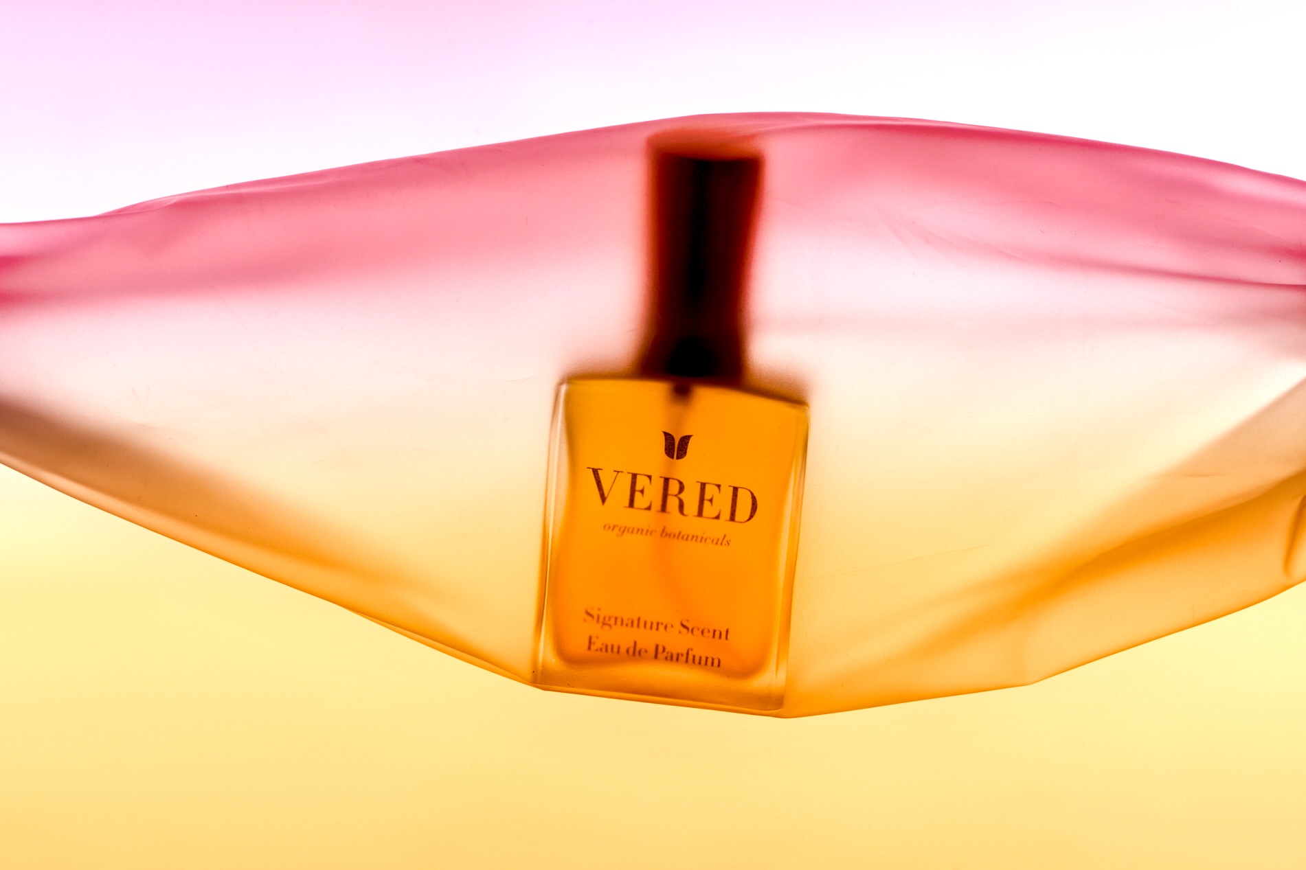 A perfume bottle labeled 'VERED' is inside a colorful, semi-transparent fabric with pink, orange, and yellow hues.