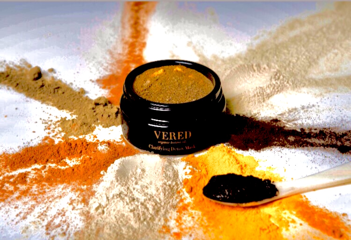 Open container of colored powder surrounded by scattered powders and a spoon on white surface.