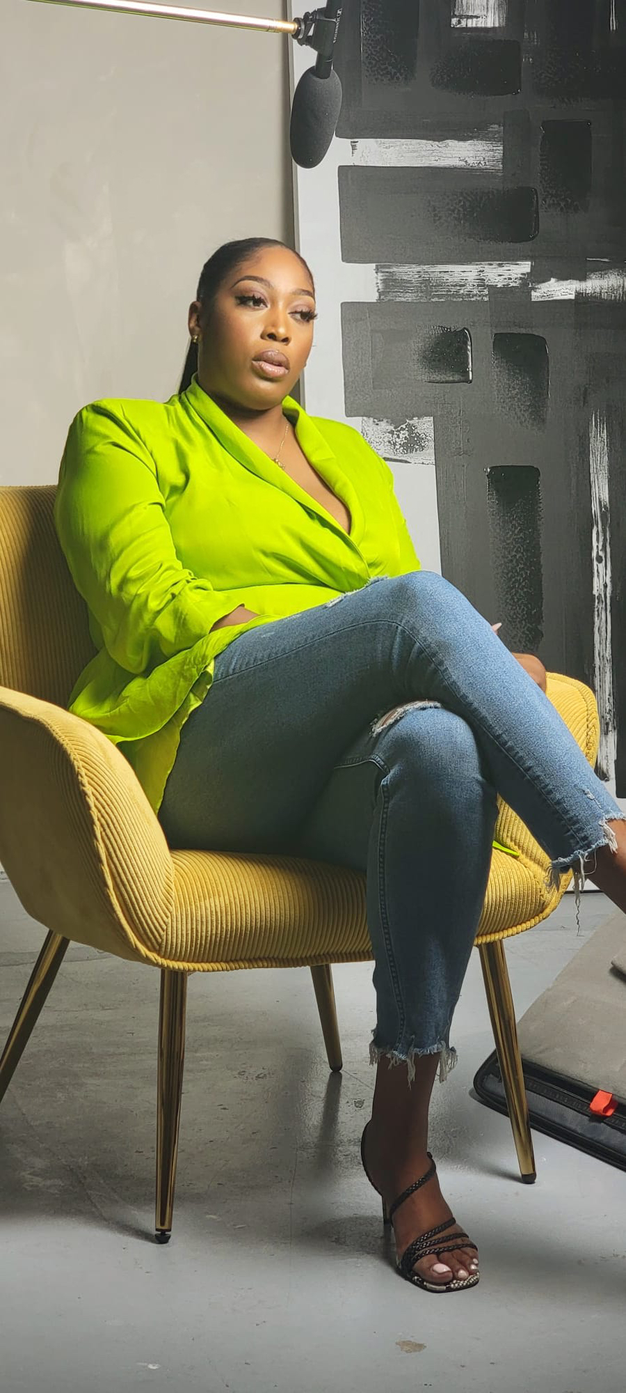 Woman sitting in a yellow chair, wearing a bright green blazer and jeans, in a modern room with black and white artwork.