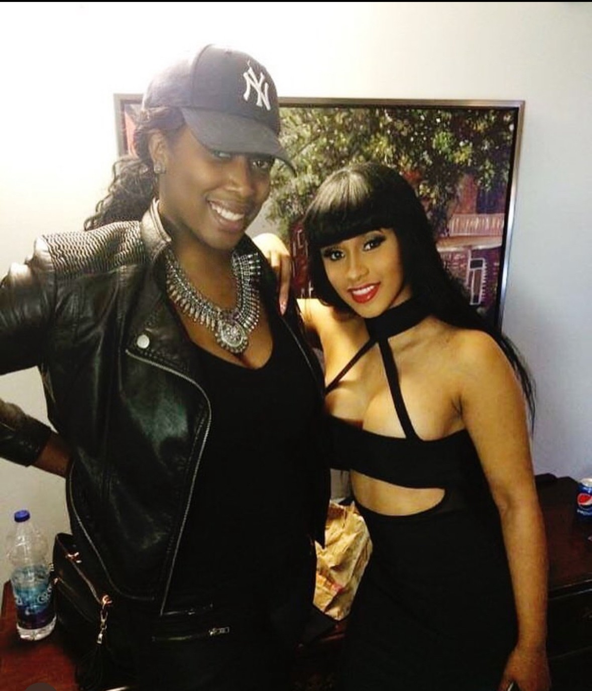 Two women smiling, one wearing a baseball cap and leather jacket, the other with straight black hair and a black outfit.