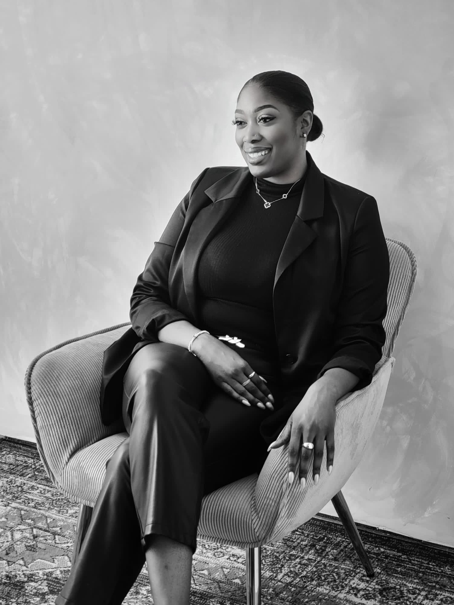 Woman sitting in a chair, smiling, wearing a blazer and pants, against a plain background.