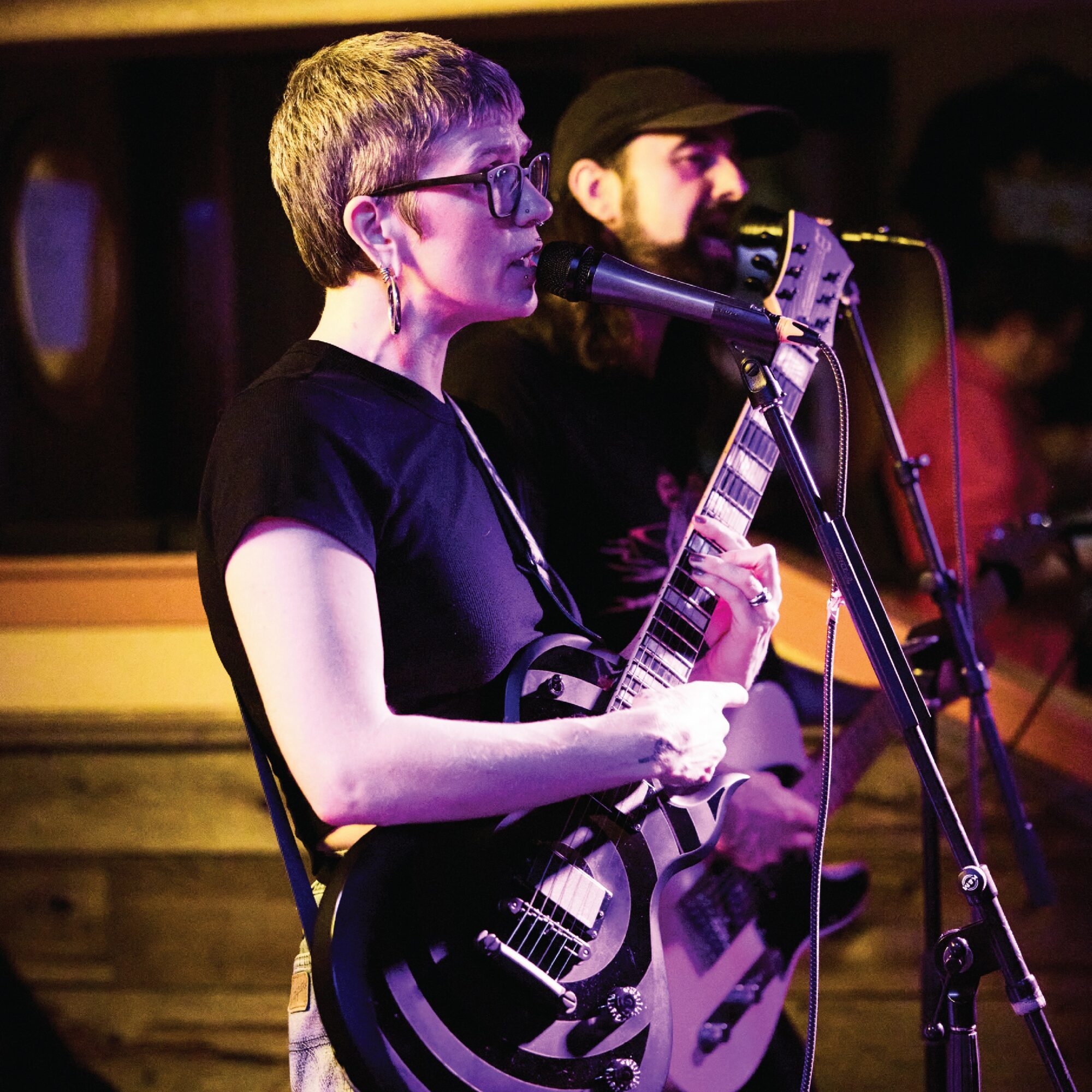 Two musicians perform on stage, one singing into a microphone and playing a guitar, the other playing guitar in background.