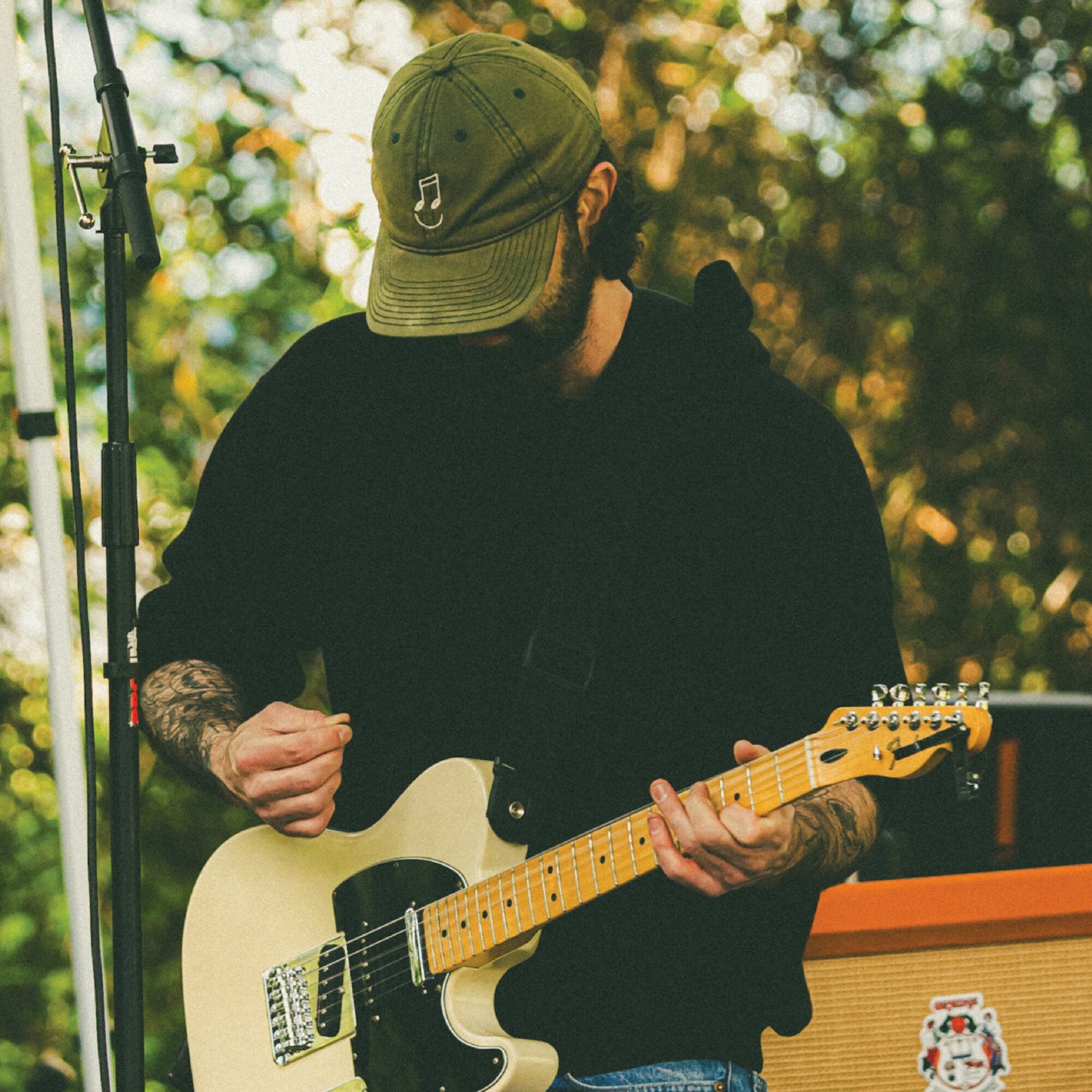 Person playing electric guitar outdoors, wearing a green cap and black hoodie, with trees in background.