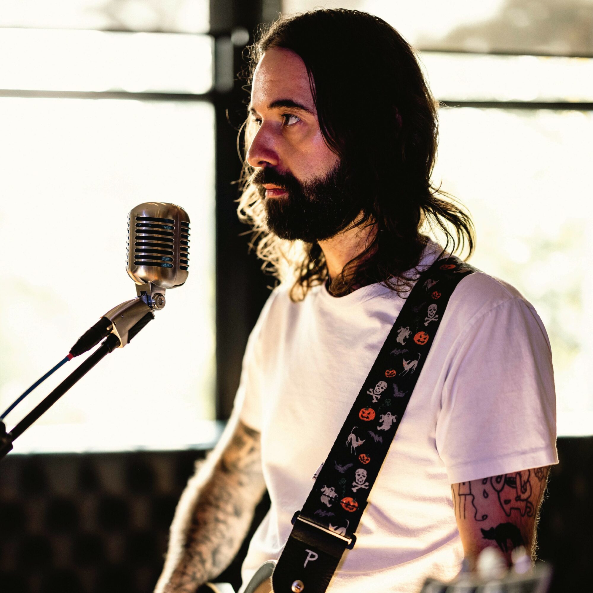 Man with long dark hair and beard singing into a microphone, wearing a white t-shirt and guitar strap.