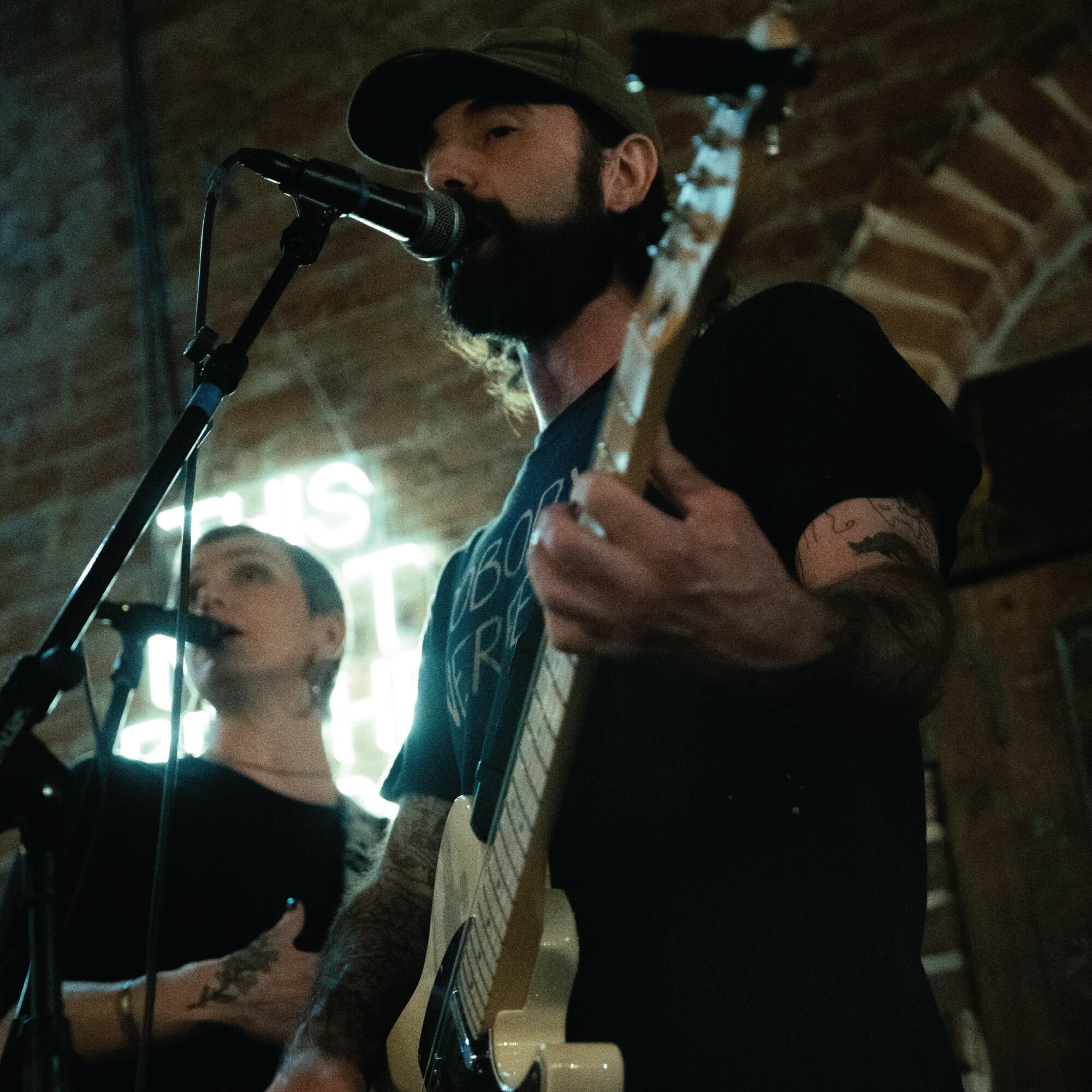 Two musicians perform with microphones and guitar in a dimly lit venue with brick walls.