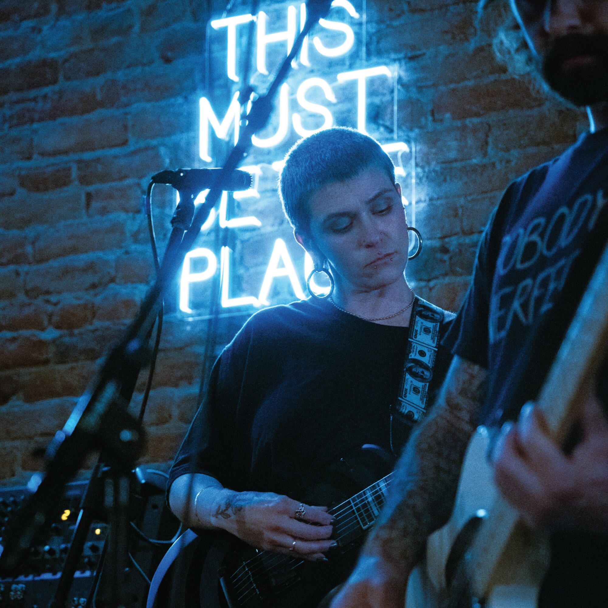 Person playing guitar on stage with a neon sign in the background reading 'This Must Be The Place'.