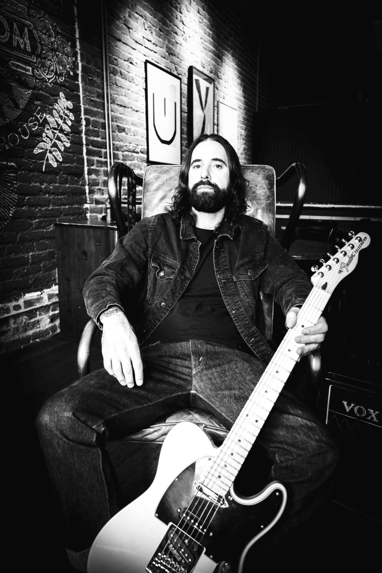 Man with beard sitting on chair holding electric guitar in a dimly lit room with brick wall background.