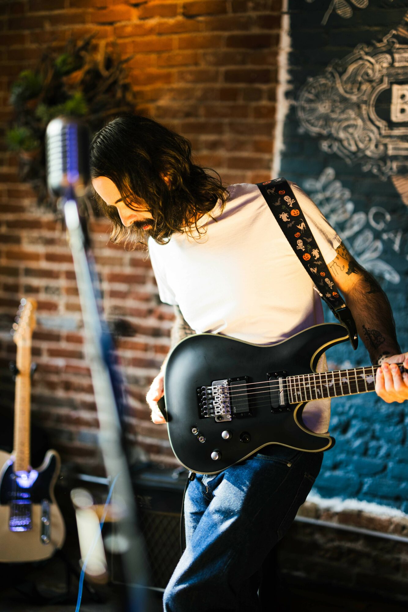 Person with dark hair playing a black electric guitar on stage with brick wall background.