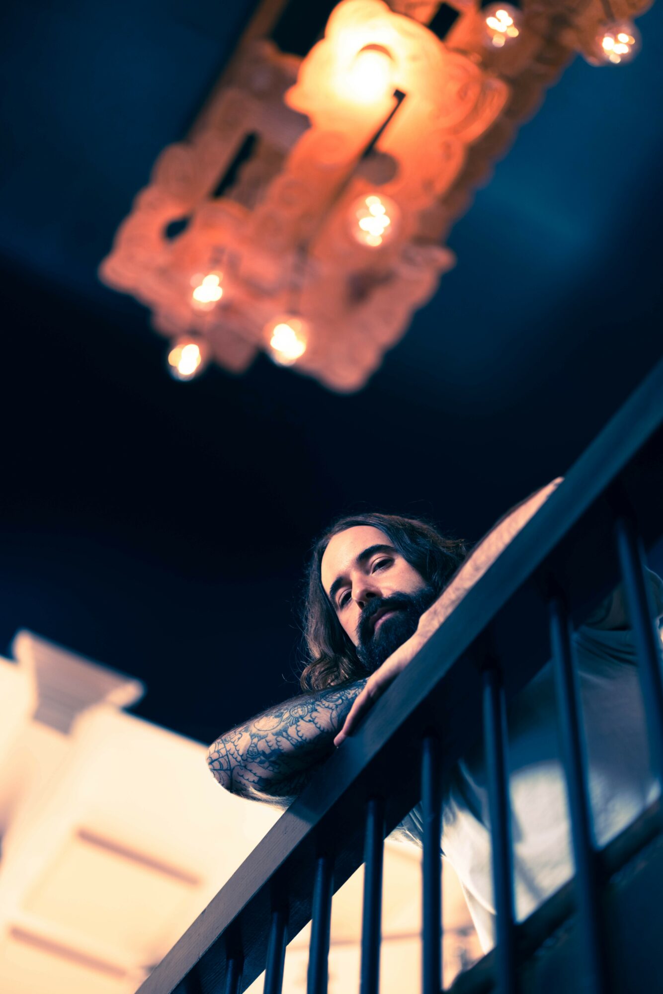Woman with dark hair and tattoos leaning on a railing indoors, chandelier hanging from ceiling above.