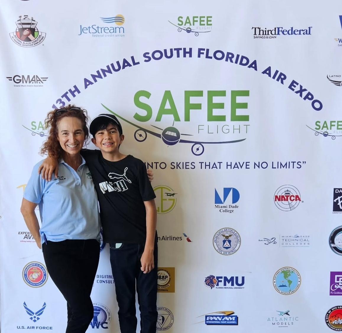 Two people smiling in front of a backdrop with event logos and text, one woman and one child, arms around each other.