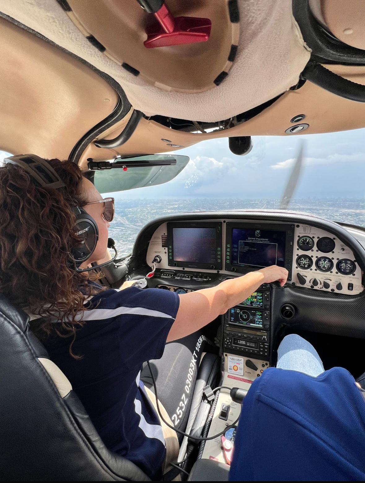 Person with curly hair wearing headphones and a dark shirt, flying a small aircraft over water and sky.