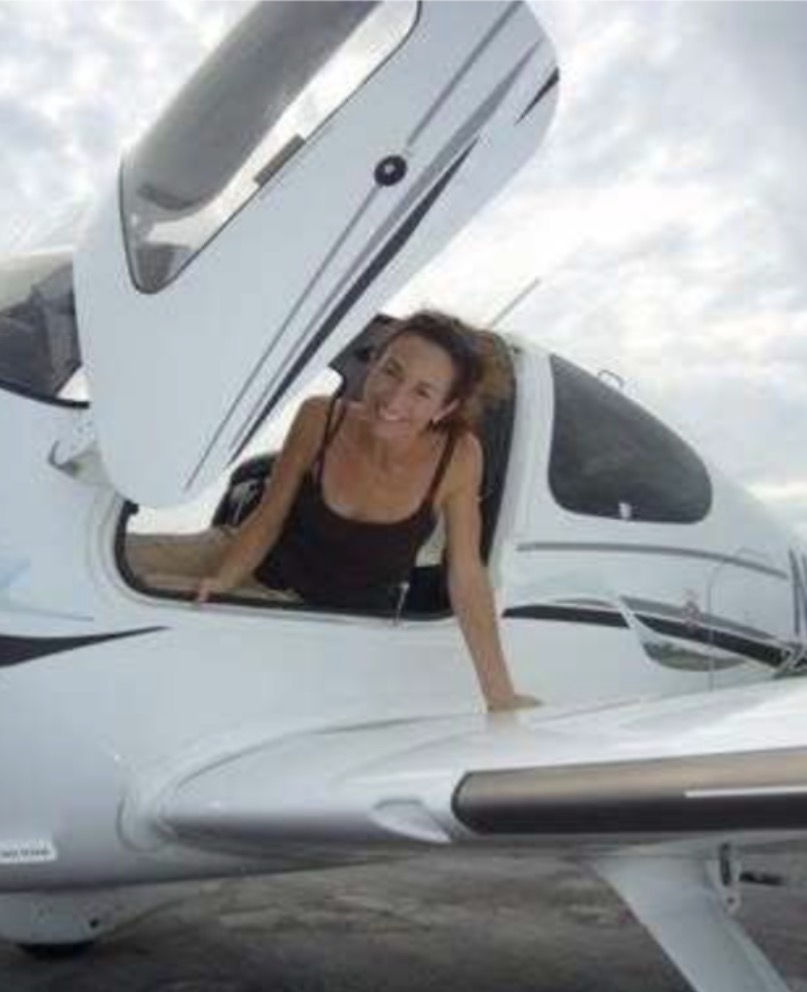 Woman leaning out of small airplane's open cockpit, smiling, with cloudy sky background.